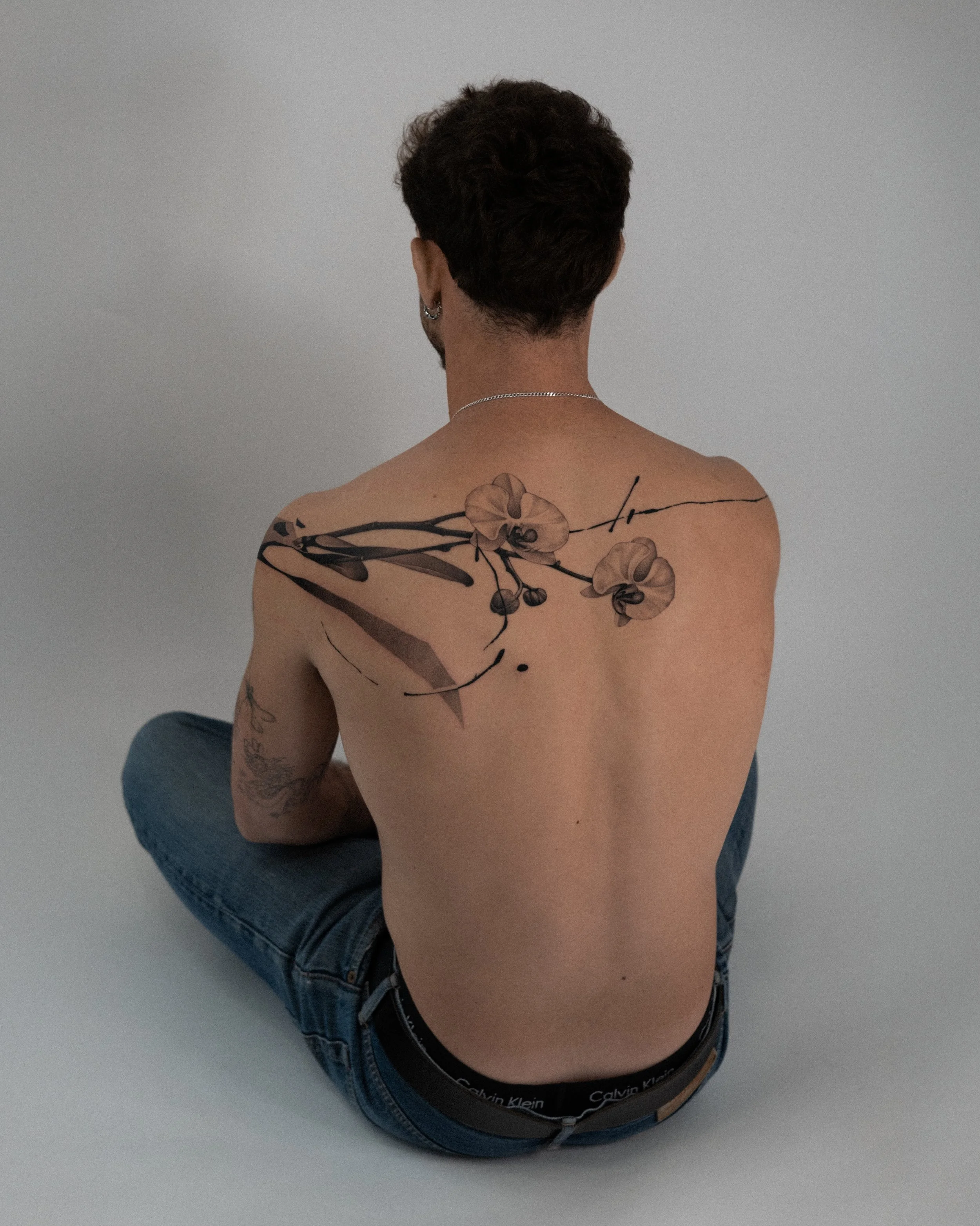 Person with short curly brown hair and tattoos on back, wearing Calvin Klein underwear and jeans, sitting against a plain white wall.