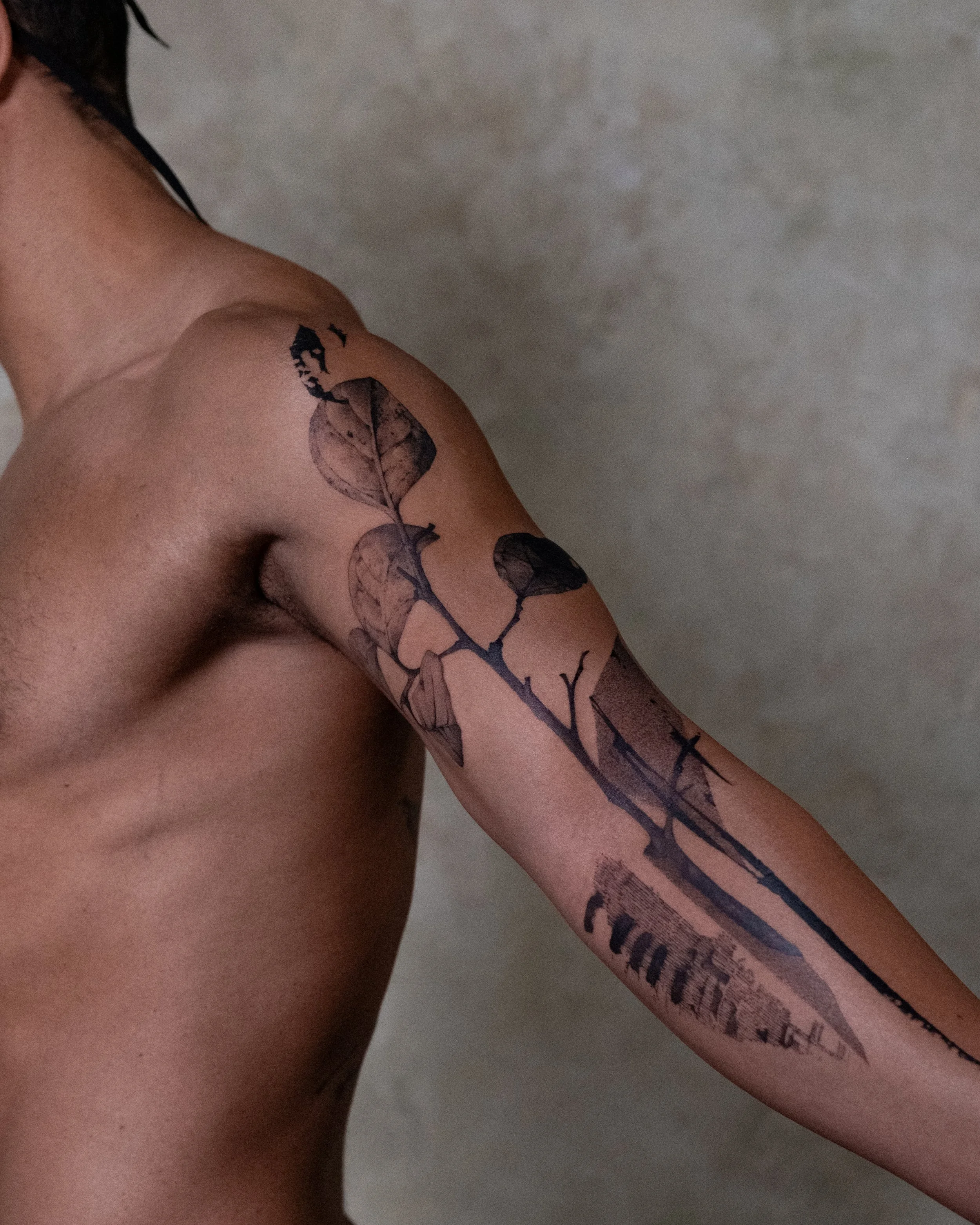Close-up of a person's shoulder and upper arm with a large tattoo of leaves and text. The background is a plain, textured wall.