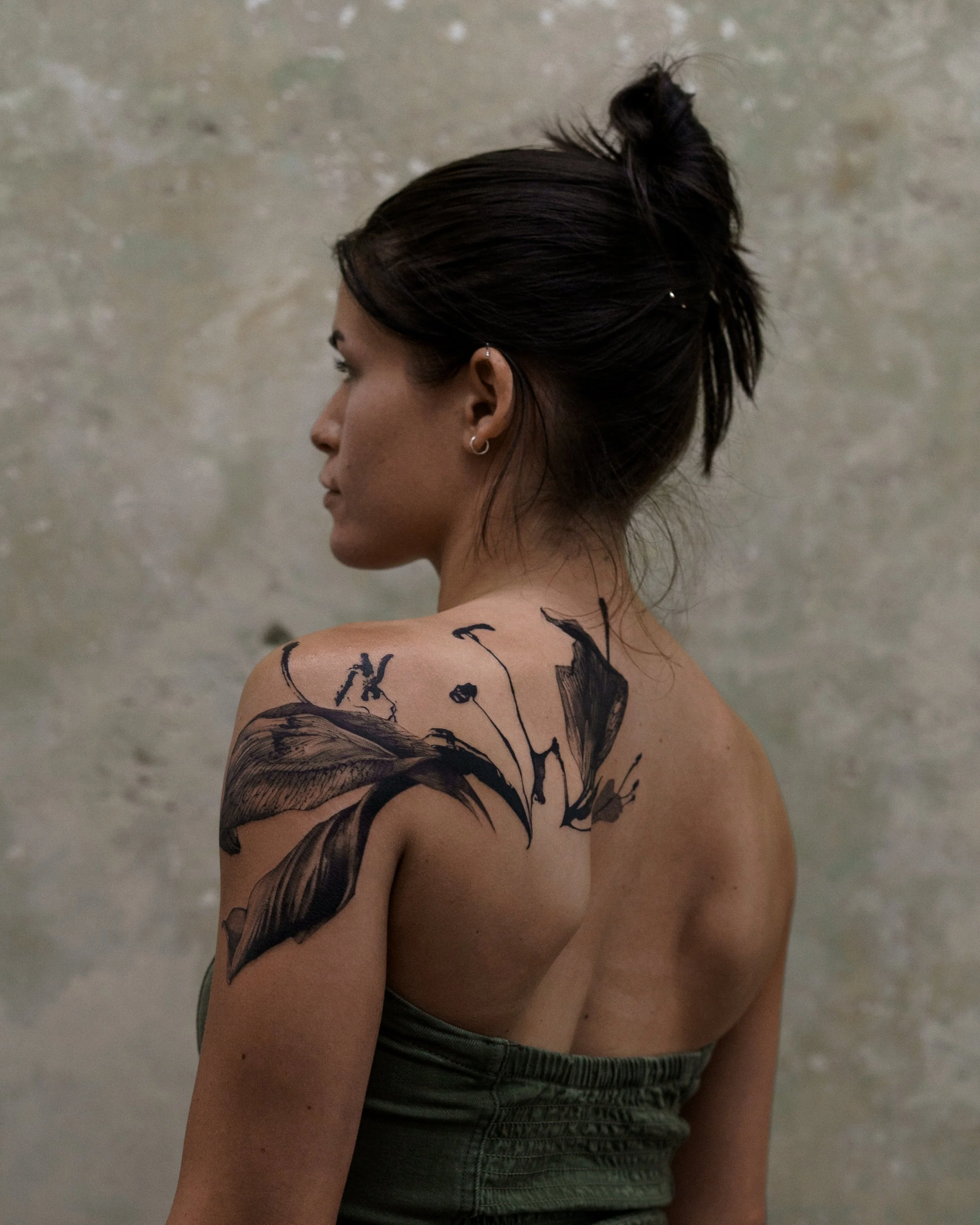 A woman with dark hair styled in a messy bun, wearing earrings, sitting with her back and side profile visible. She has a tattoo on her arm and shoulder and is wearing dark green shorts. The background is a plain, textured wall.
