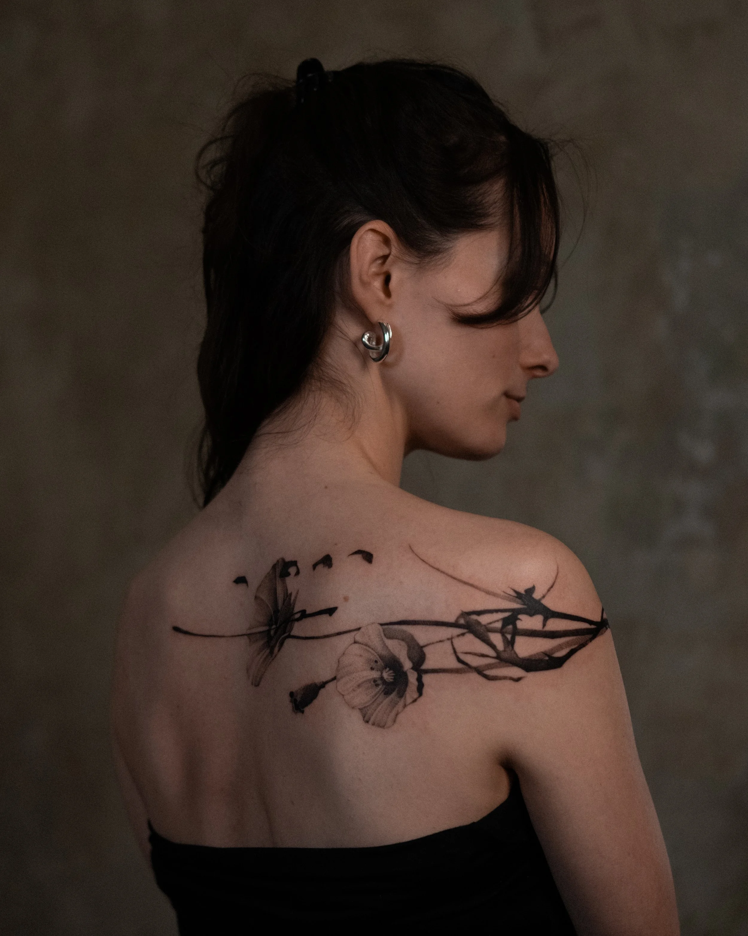 Close-up of a woman with tattoos, silver hoop earrings, dark hair, and wearing a black top, posed with her face turned to the side, against a dark background.