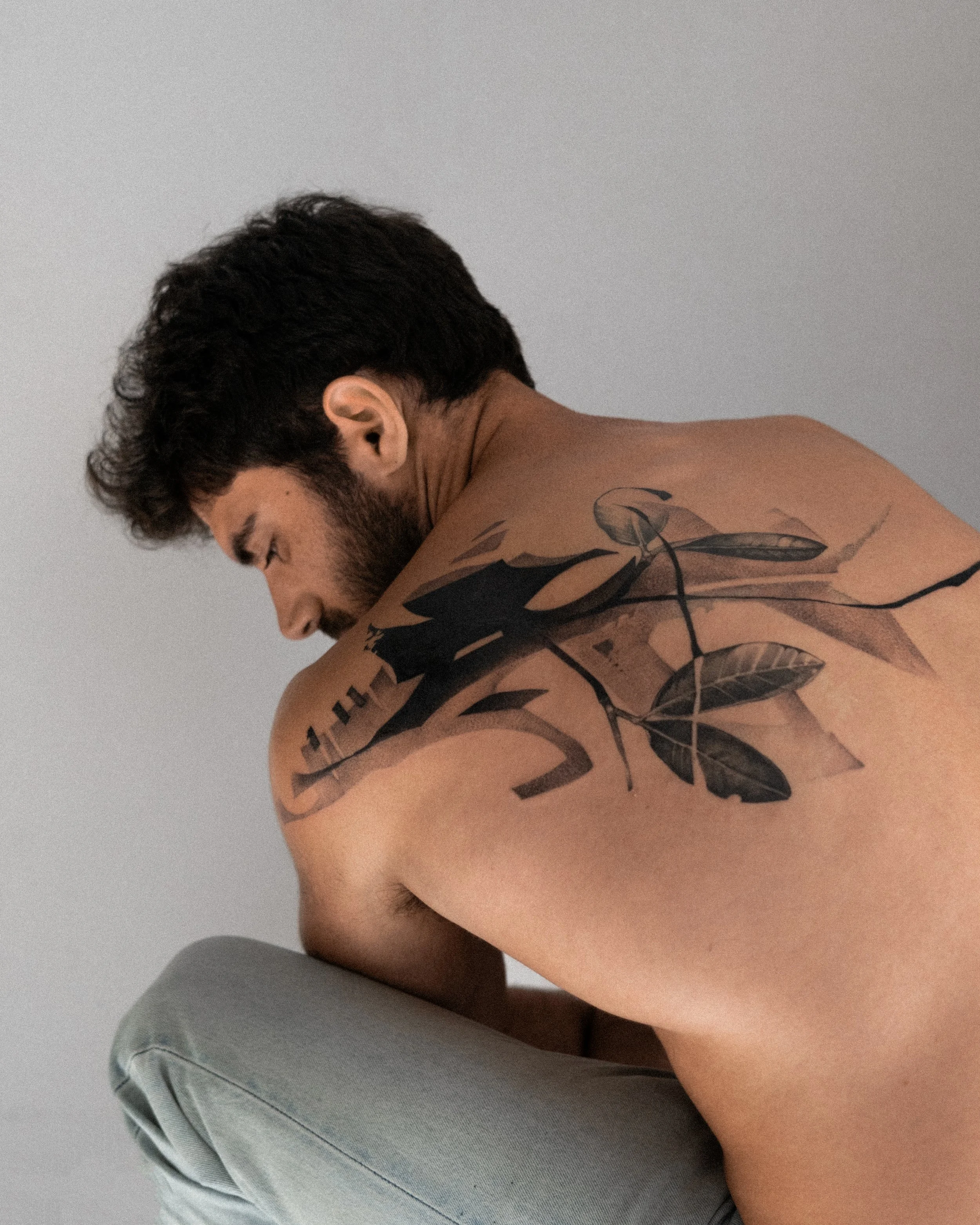 A shirtless man with dark hair and a beard, sitting against a plain gray wall, with tattoos on his shoulder and upper arm.