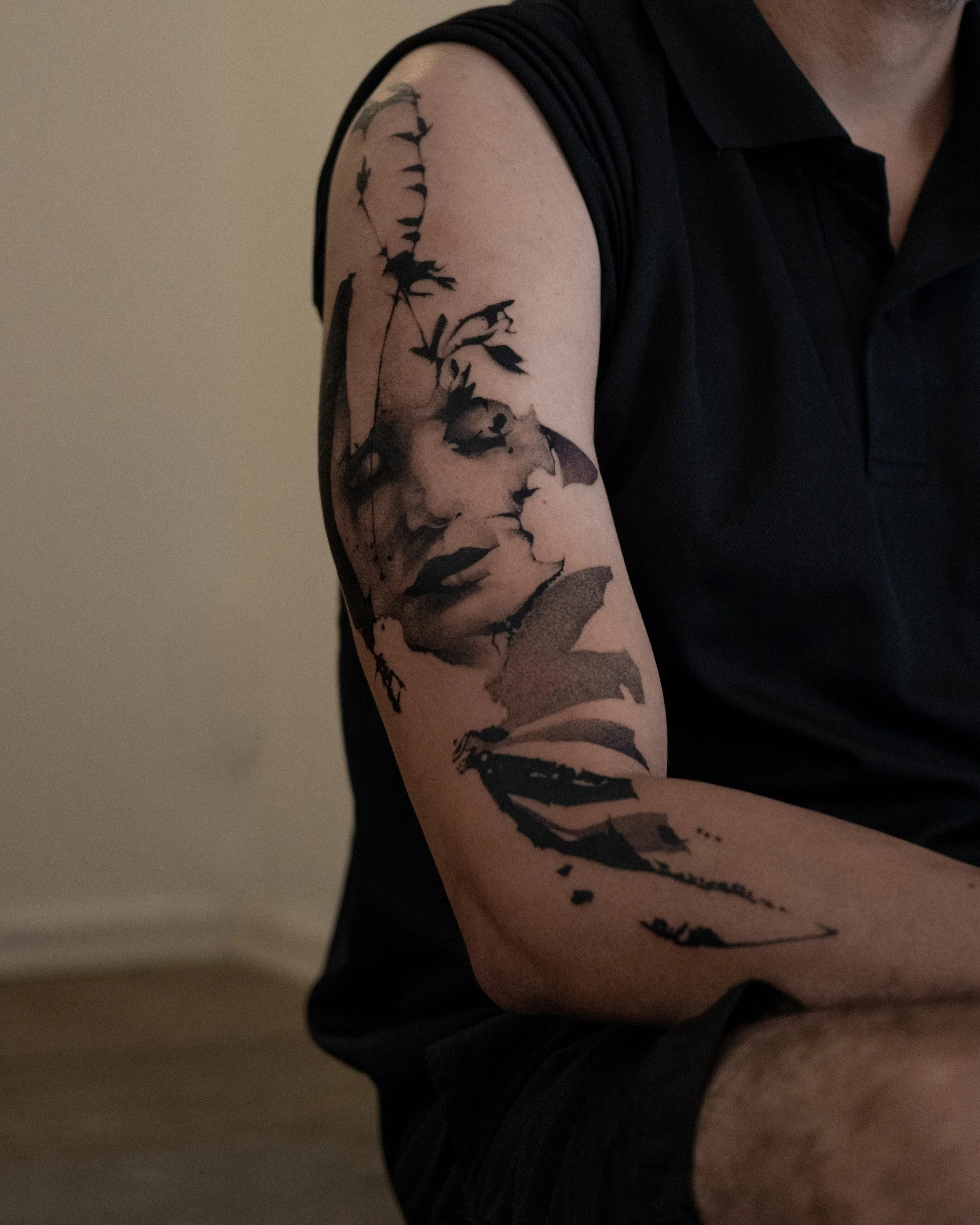 A person wearing a black short-sleeve shirt displaying a large tattoo on their upper arm. The tattoo features a highly detailed portrait of a woman with flowers and foliage around her face, rendered in black ink.
