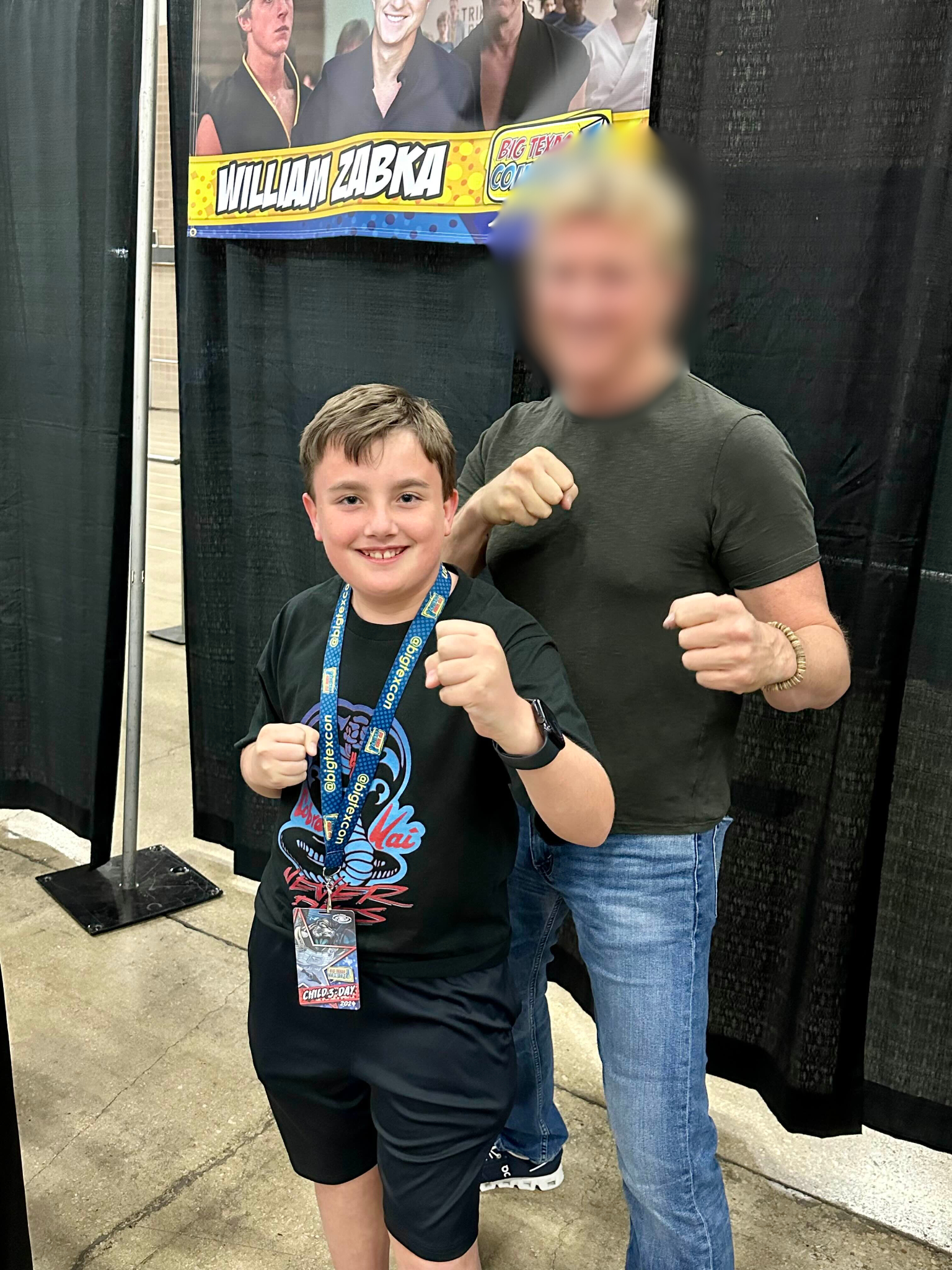 A young boy and an adult man posing with their fists raised at a comic convention. The boy is wearing a black T-shirt with a graphic design, a lanyard with a badge, and a smartwatch. The man is wearing a dark T-shirt and jeans. Behind them, there is a black curtain and a poster with images of women and the name William Zabka.