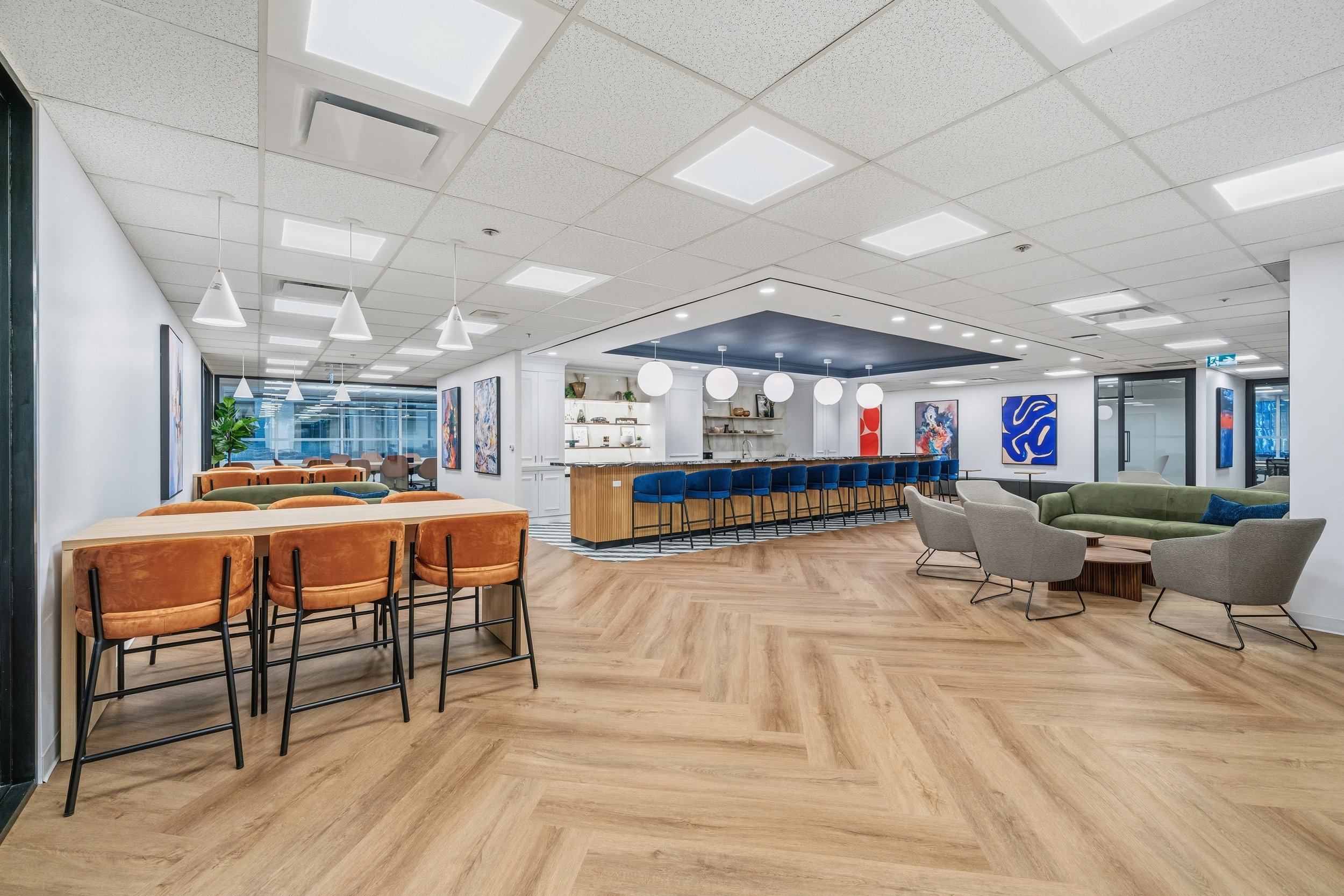 Modern office lounge with seating areas, abstract art on the walls, a bar with barstools, and a bookshelf in the background.