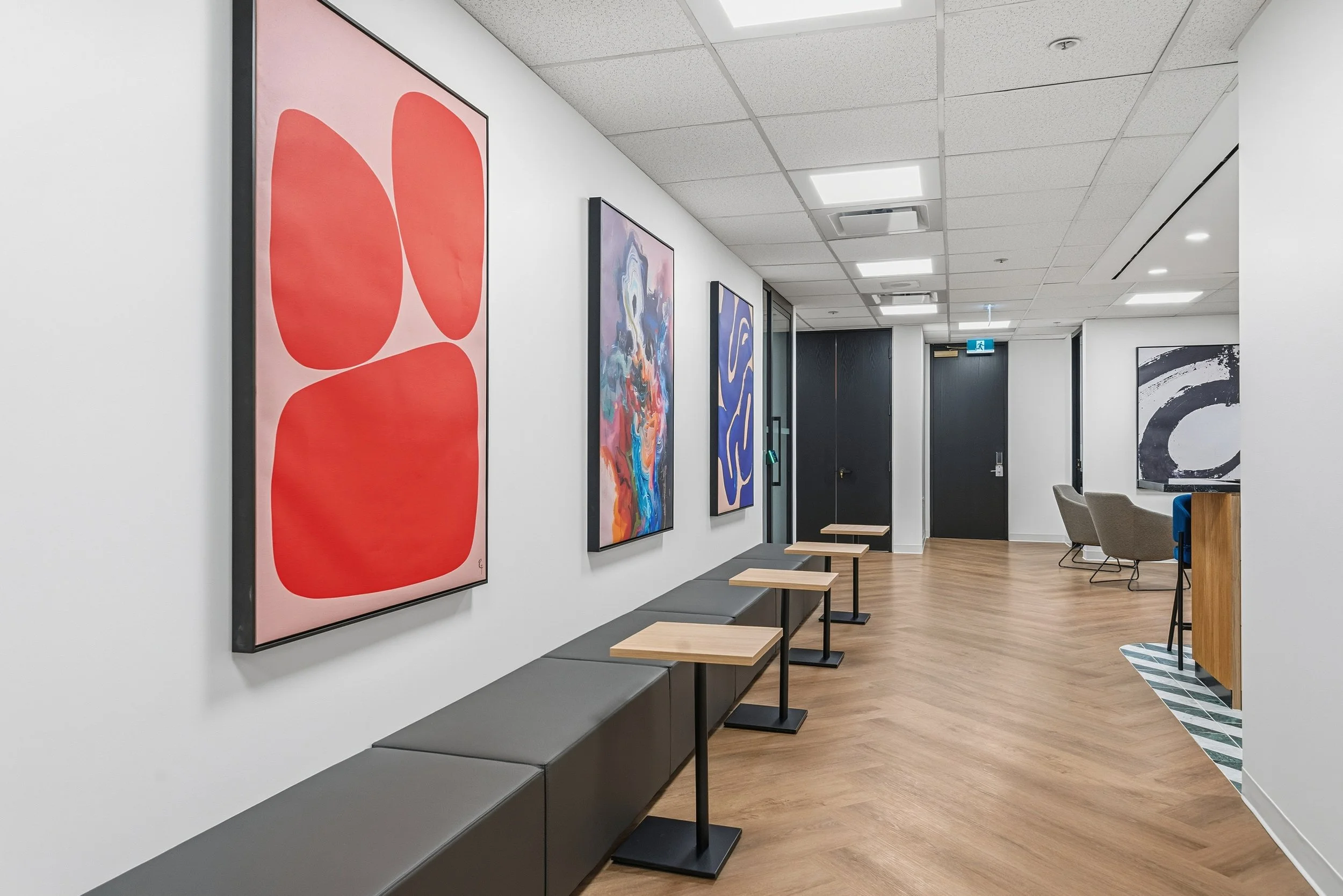 Modern waiting area with abstract art artwork on the white walls, small tables, and chairs with wood and gray upholstery, in a brightly lit, clean space.