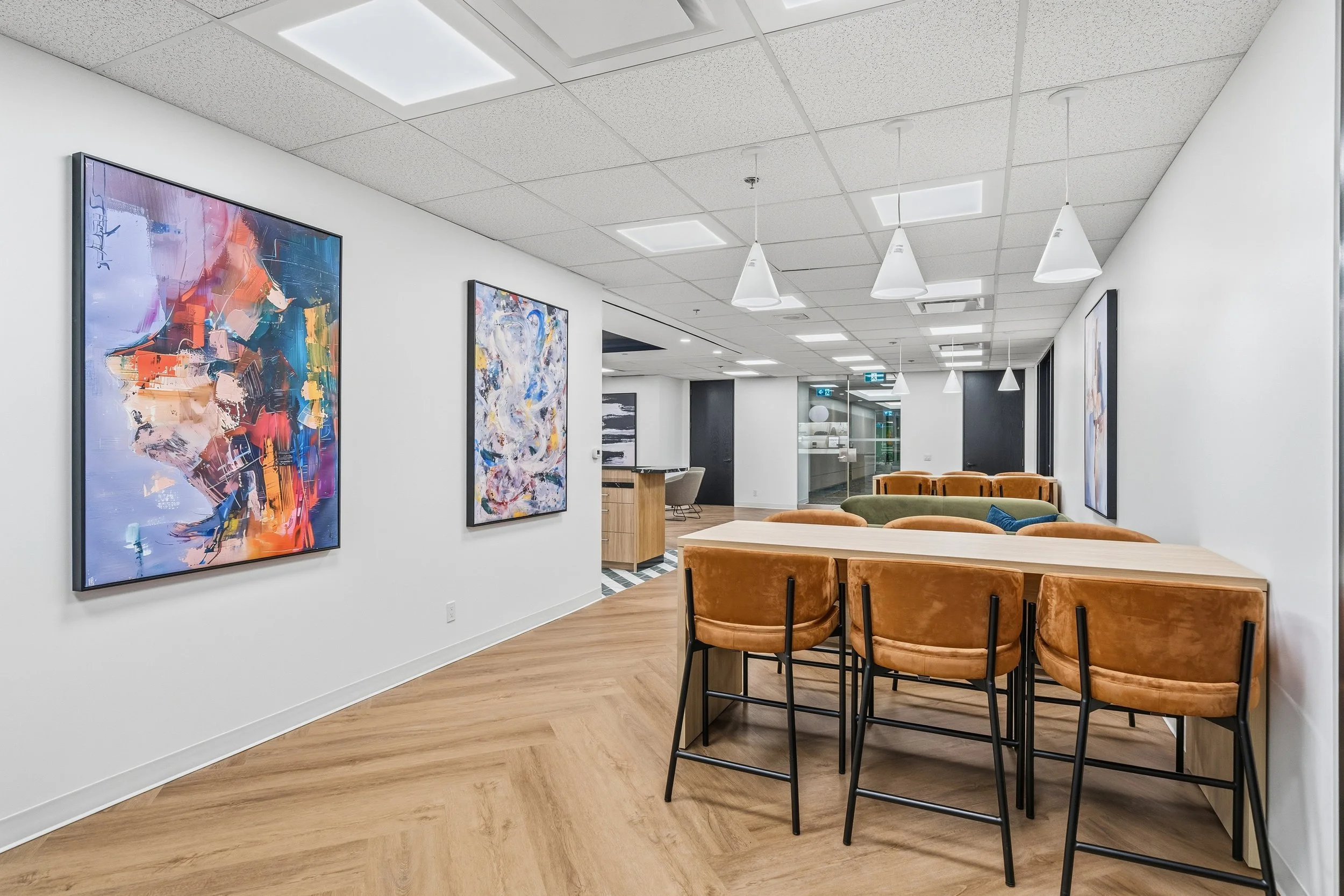 Modern office lounge area with abstract art paintings on white walls, a long wooden table surrounded by orange velvet chairs, wooden flooring, and contemporary pendant lighting.