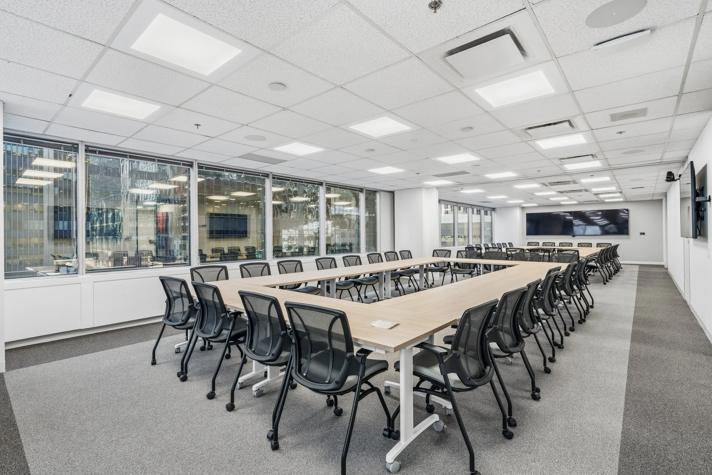 Large empty conference room with U-shaped table setup, black mesh chairs, large windows on one side, and two wall-mounted monitors at the front.
