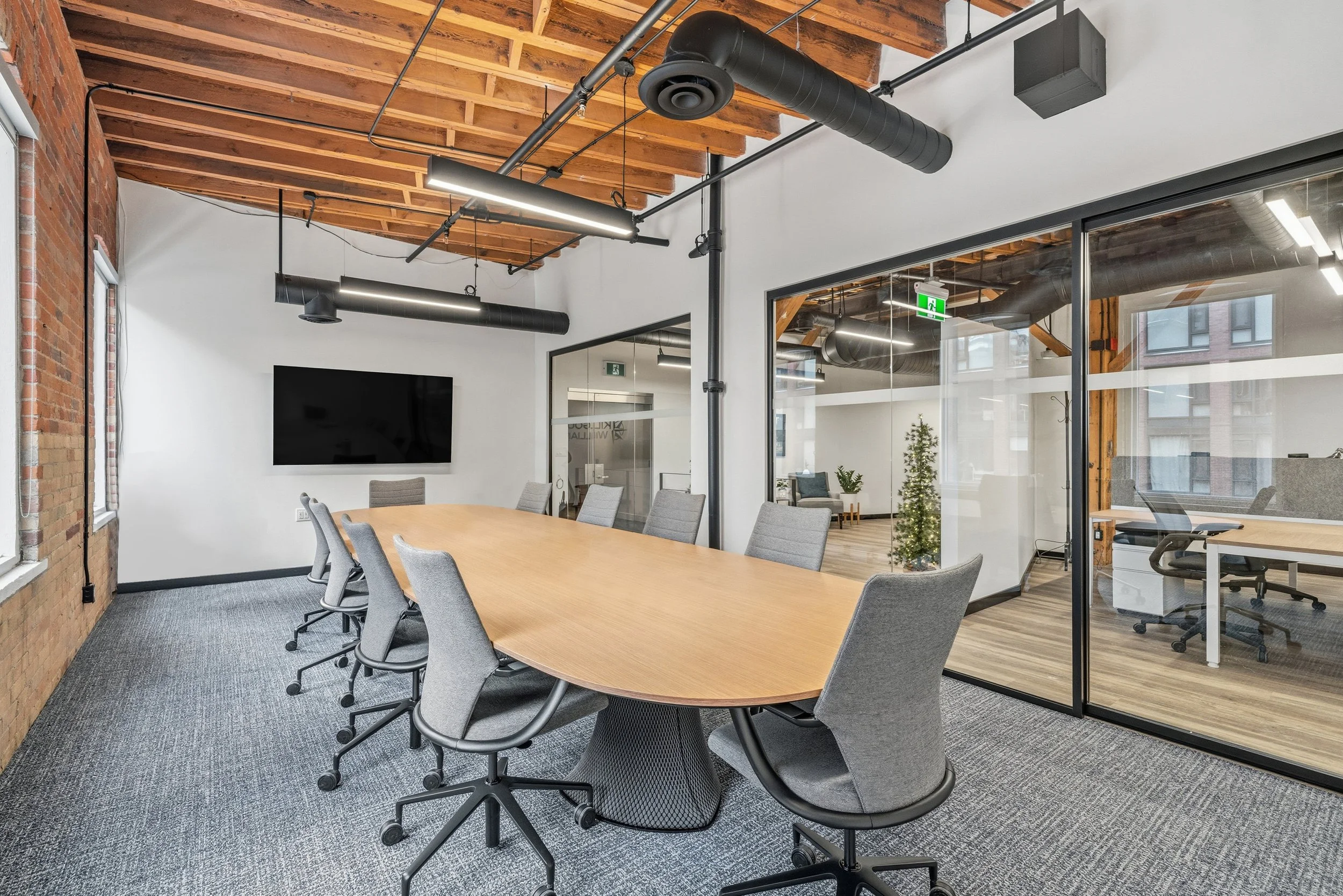 Conference room with a large wooden table and gray office chairs, brick wall, large window, mounted TV, ceiling with exposed wooden beams and black industrial ducts, glass partition separating another office space with a Christmas tree.