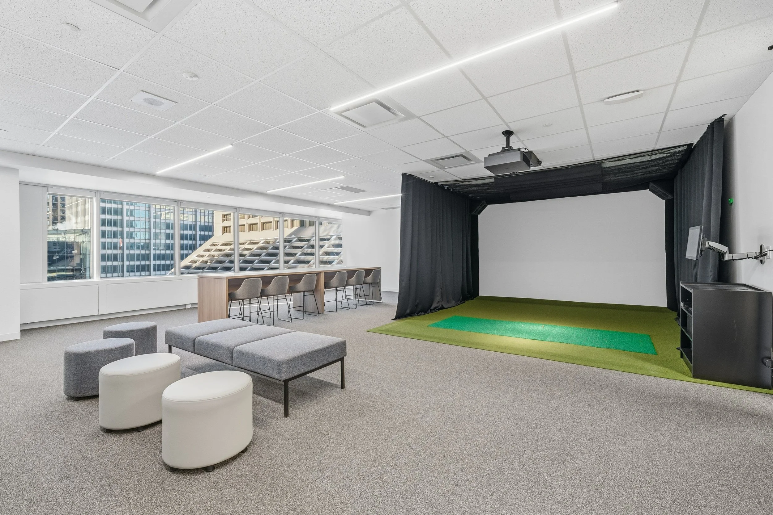 Modern office or conference room with a seating area, large windows, and a small presentation or golf simulation area with a green mat and black curtains.