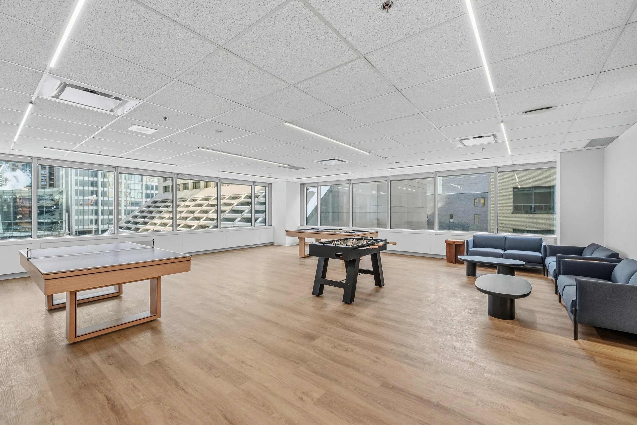 Empty recreational room with ping pong table, foosball table, sitting area with sofas, and large windows showing cityscape outside.