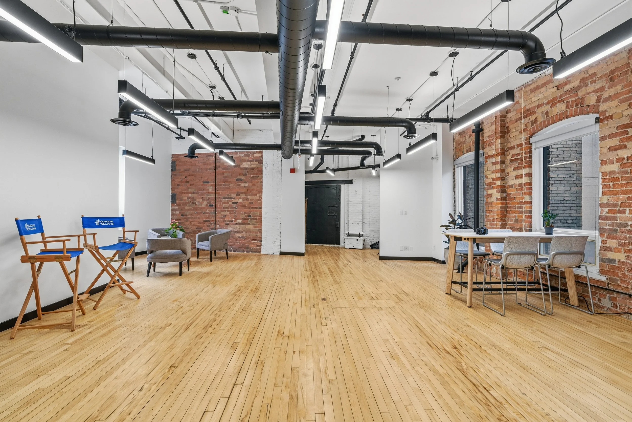 Empty room with wooden floor and exposed brick and white walls, black ceiling ducts, windows, and modern furniture including chairs and a table, with lighting fixtures hanging from the ceiling.