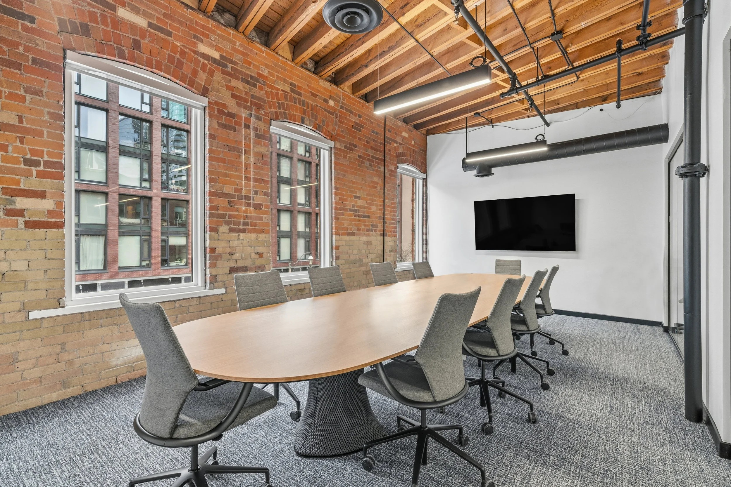 Conference room with a long wooden table, nine gray office chairs, exposed brick walls, large windows, a mounted flat screen on a white wall, and an industrial ceiling with wooden beams, metal pipes, and modern linear lighting.