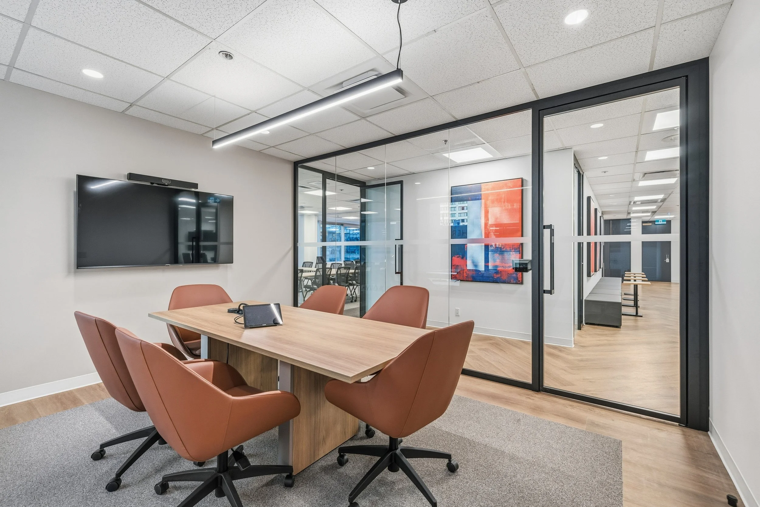 Modern conference room with a wooden table, six brown leather chairs, a wall-mounted screen, and glass walls in an office setting.
