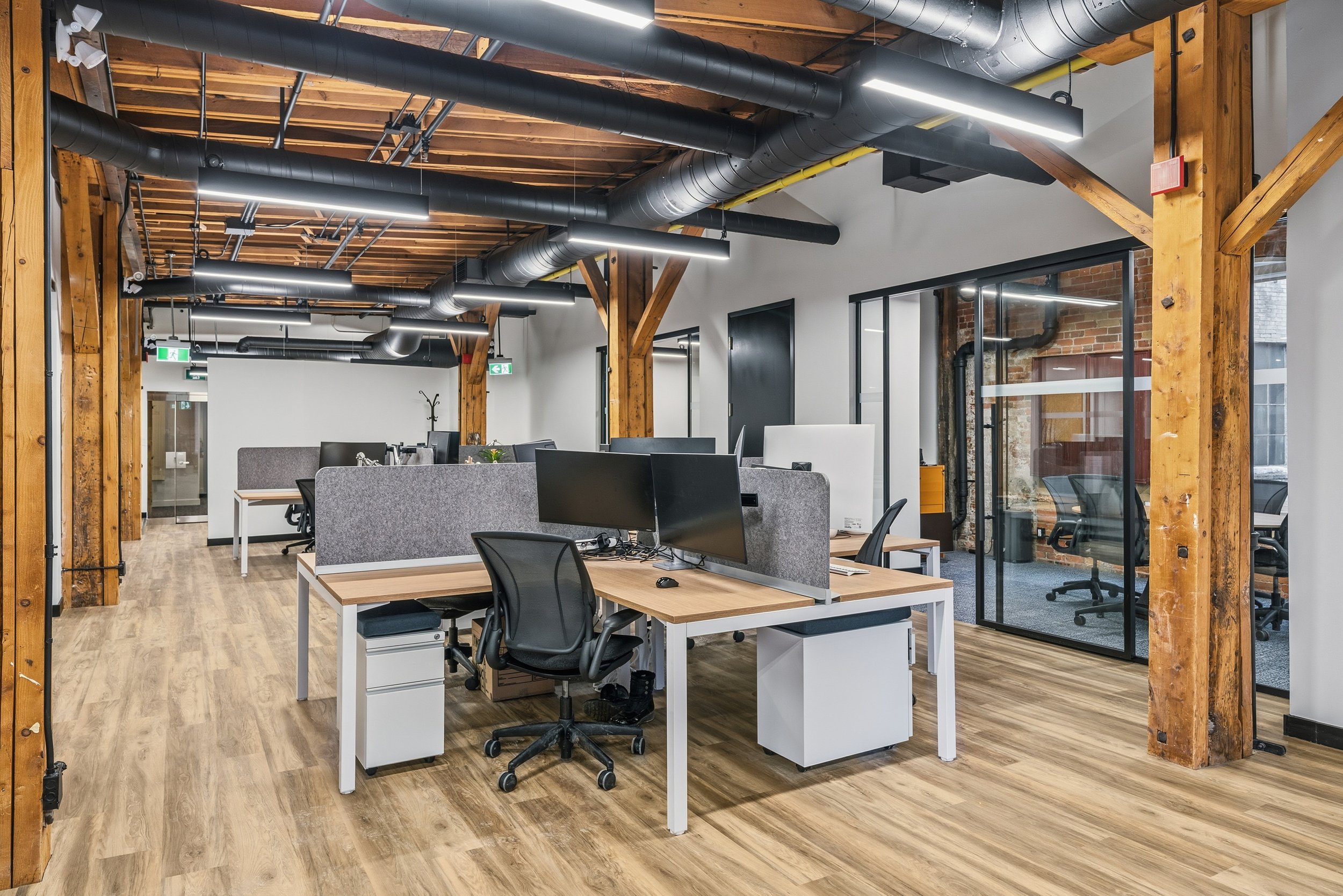 Modern office space with wooden beams, exposed ductwork, and multiple desks with computer monitors and chairs.