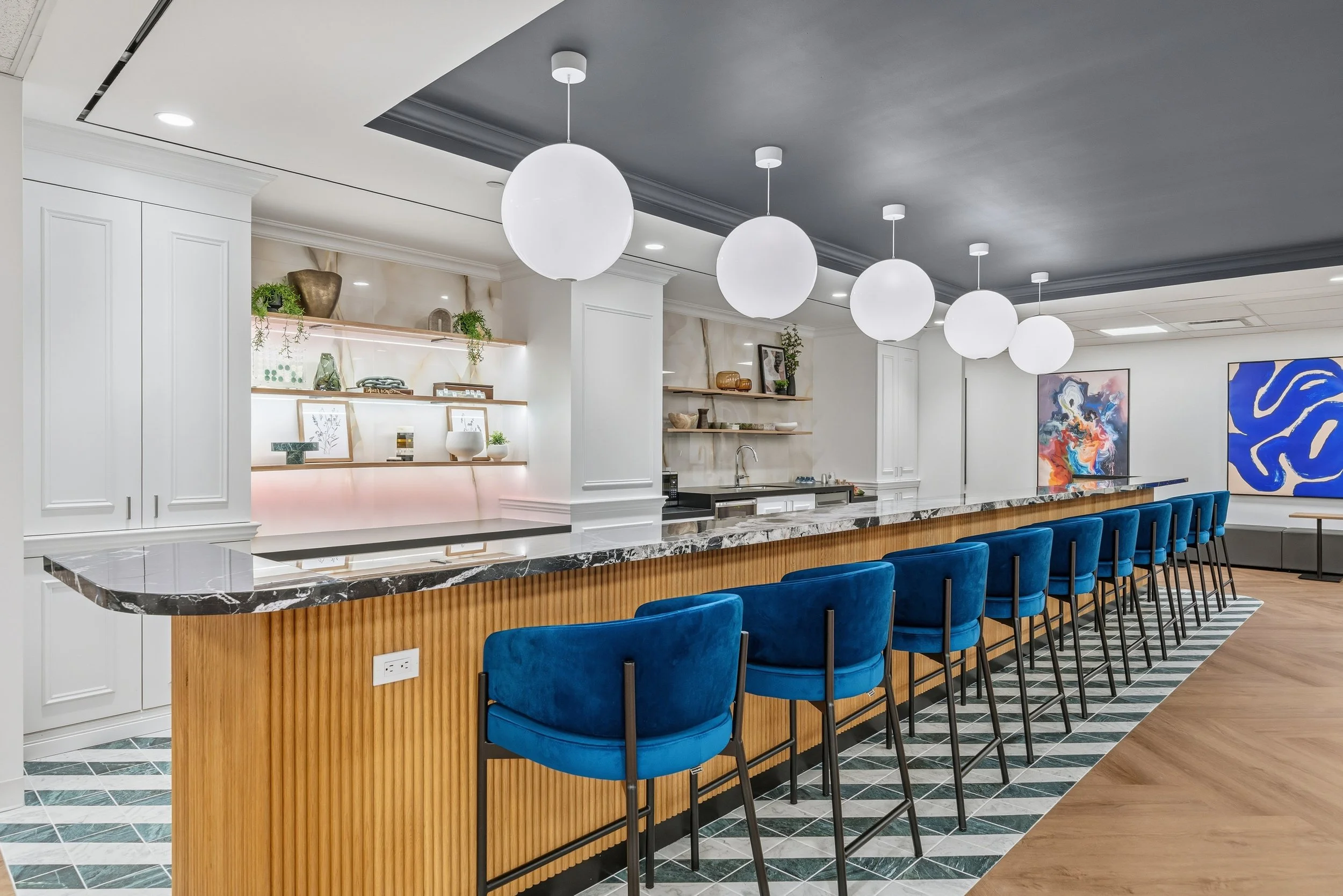 Modern bar with a marble countertop, blue velvet chairs, and artwork on the walls.