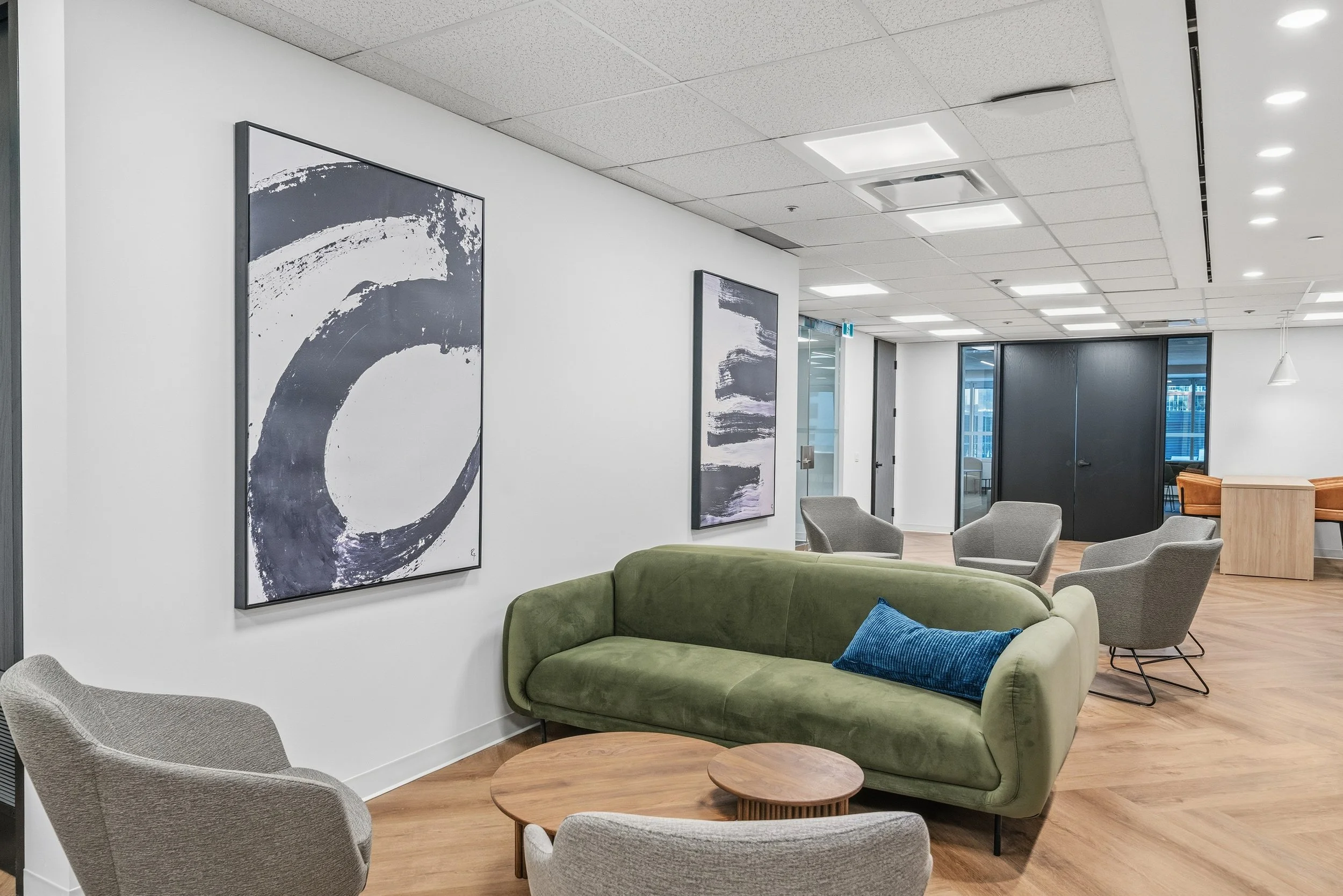 Modern office lounge with green sofa, gray chairs, wooden coffee tables, abstract artwork on white walls, large windows, and ceiling lights.