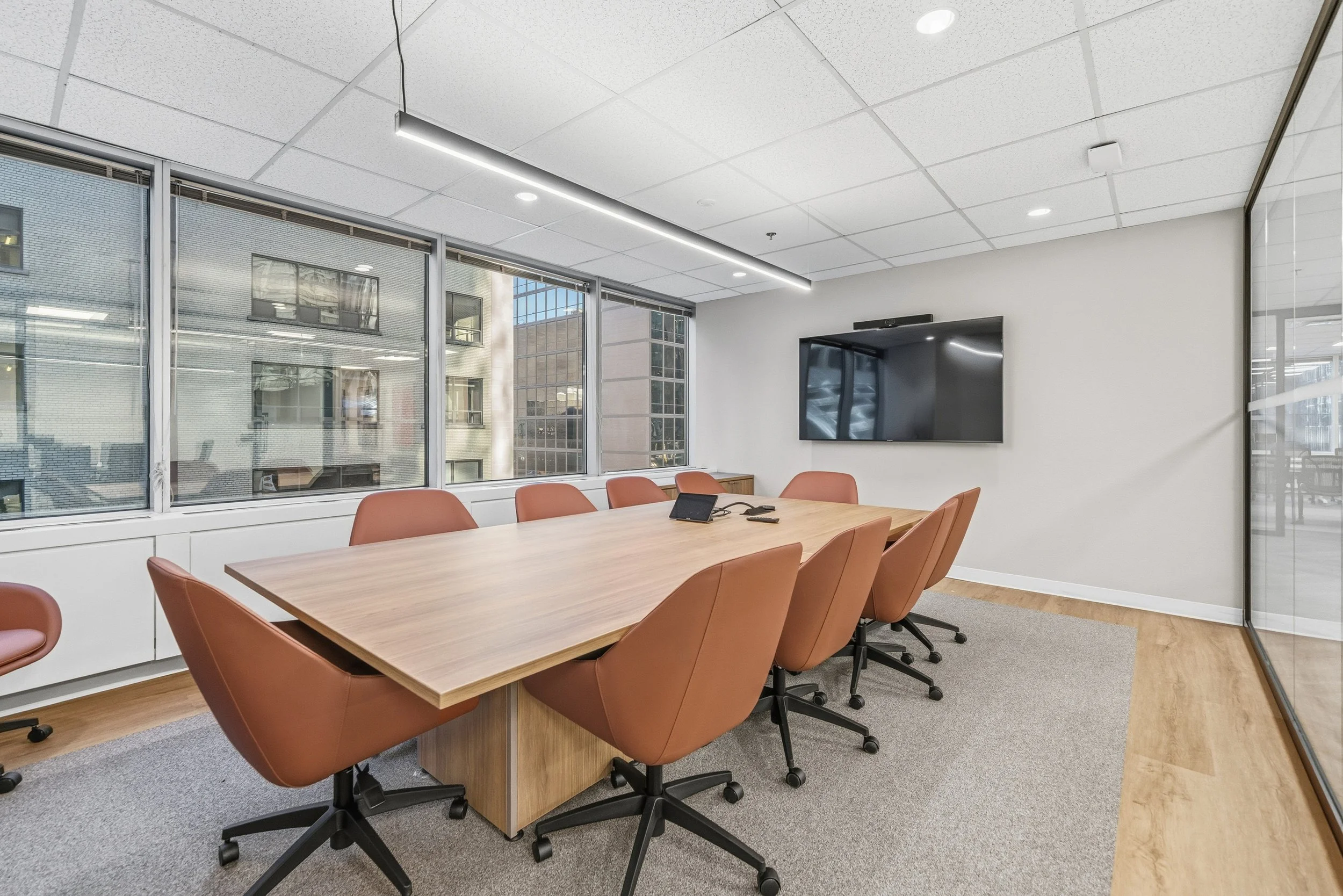 Modern conference room with a large wooden table, eight burnt orange chairs, a flat-screen TV on the wall, large windows with city view, and a glass wall on the right side.