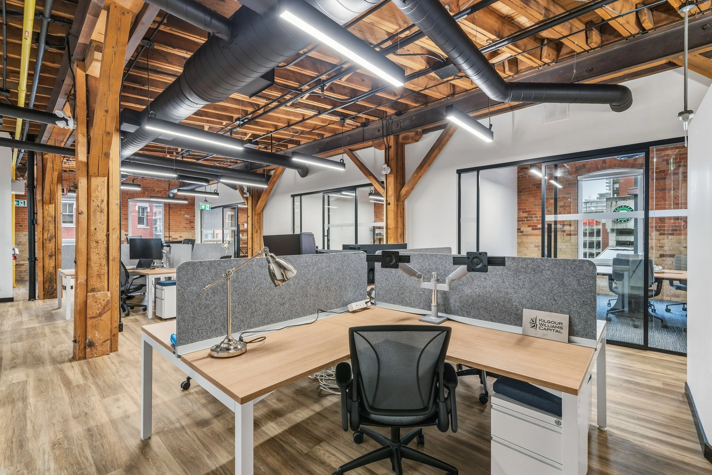 Modern open-plan office with multiple workstations, grey partition panels, wooden beams, exposed ductwork, and large windows with brick buildings outside.