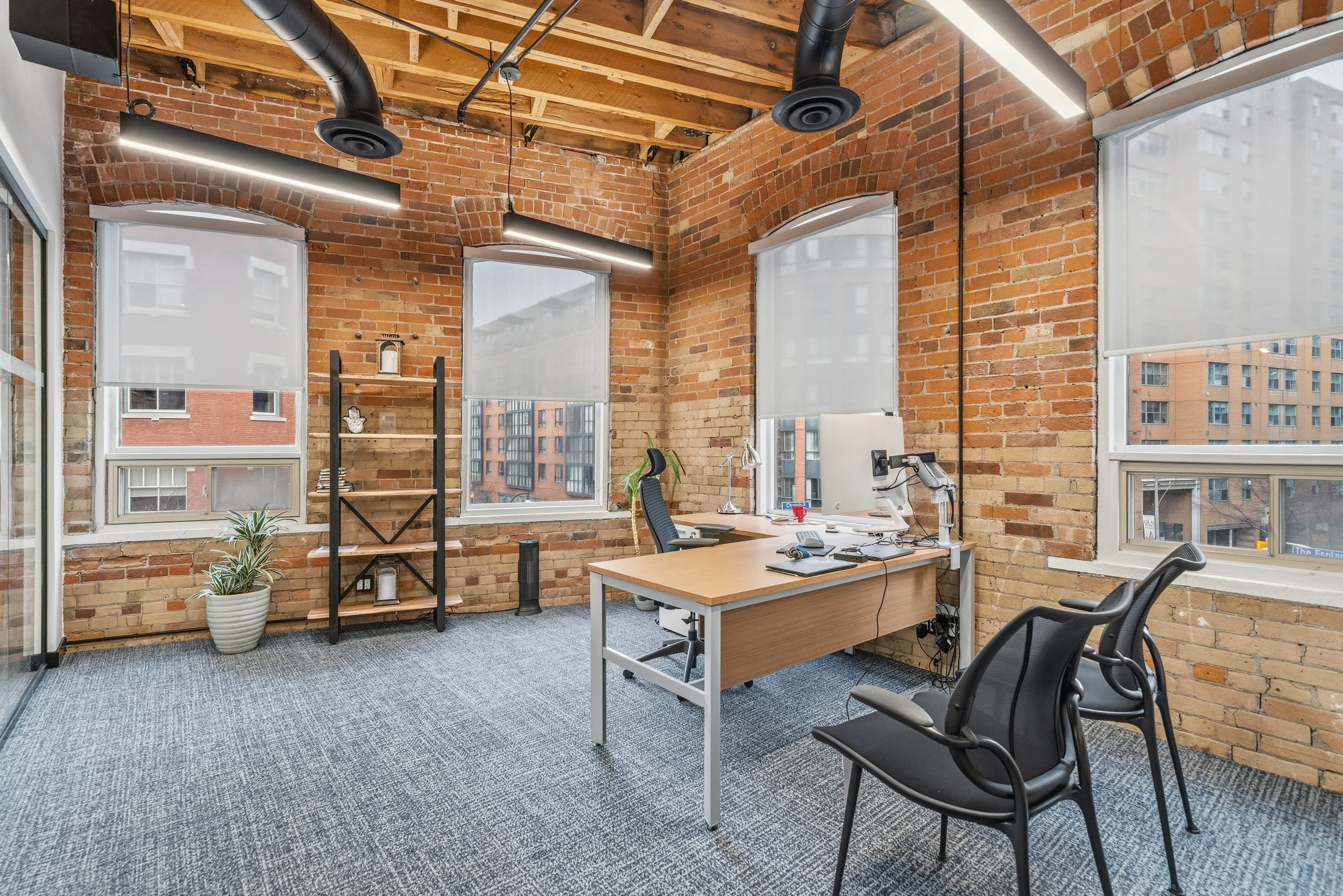 An office with exposed brick walls, large windows with white blinds, and industrial-style lighting. There is a wooden desk with a computer, monitor, and office supplies, along with a black swivel chair and two black guest chairs. A metal shelf with d