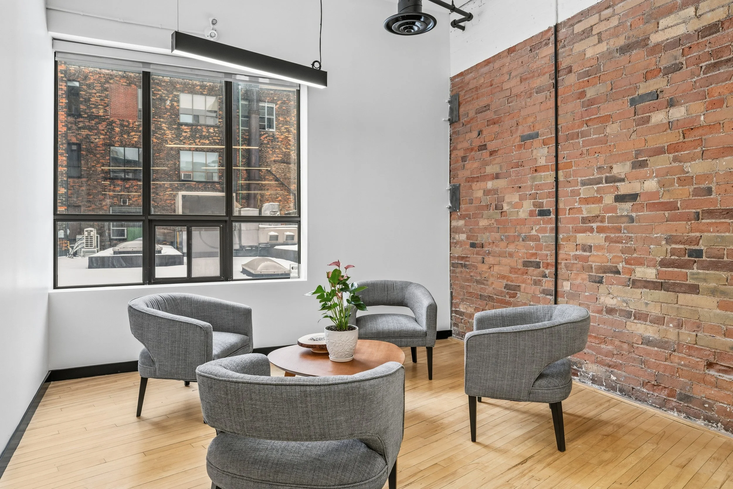 Interior of a modern office or lounge area with four gray armchairs arranged around a small round wooden table with a potted plant, large window with black framing, and exposed brick wall.
