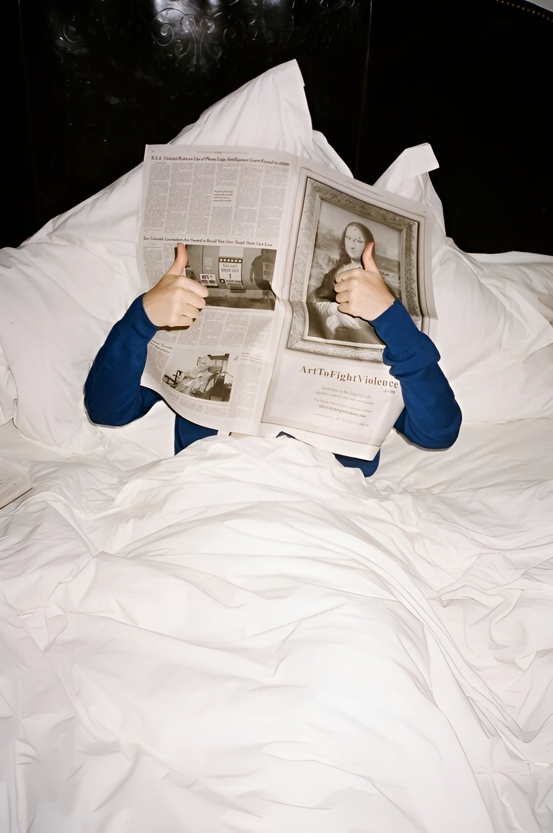 Person lying in bed, covered with white sheets, reading a newspaper with some articles and images, and holding a magazine open with an artwork titled 'Art of Fight Violence' and a portrait of a woman.
