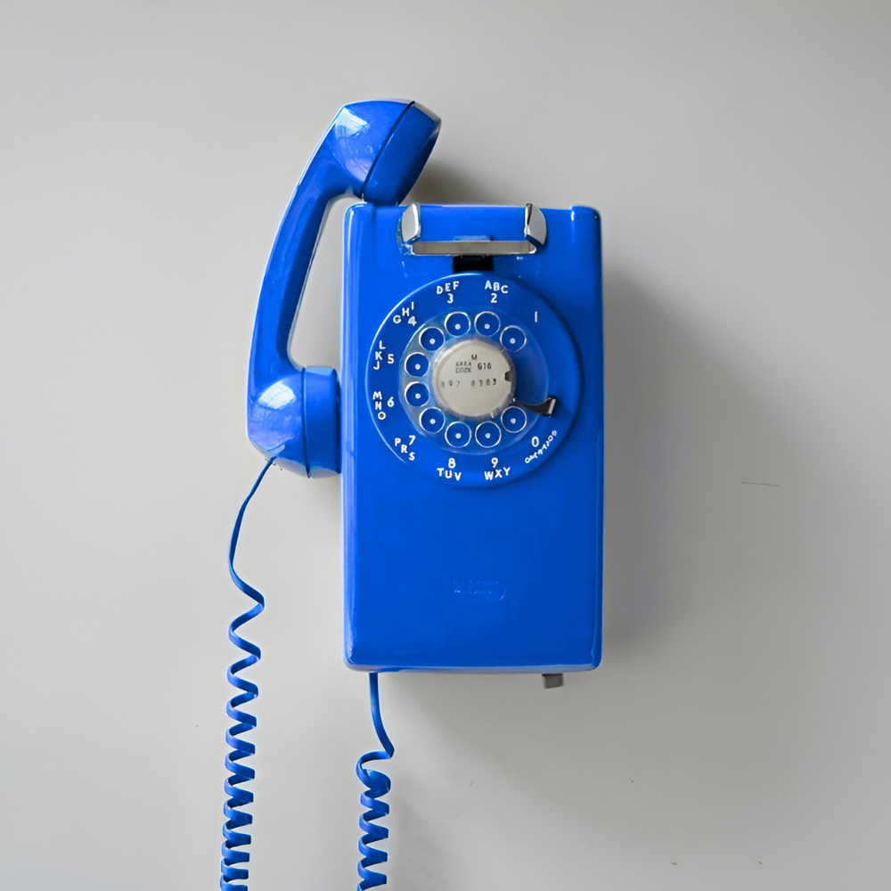 A vintage blue rotary dial telephone mounted on a plain light gray wall.