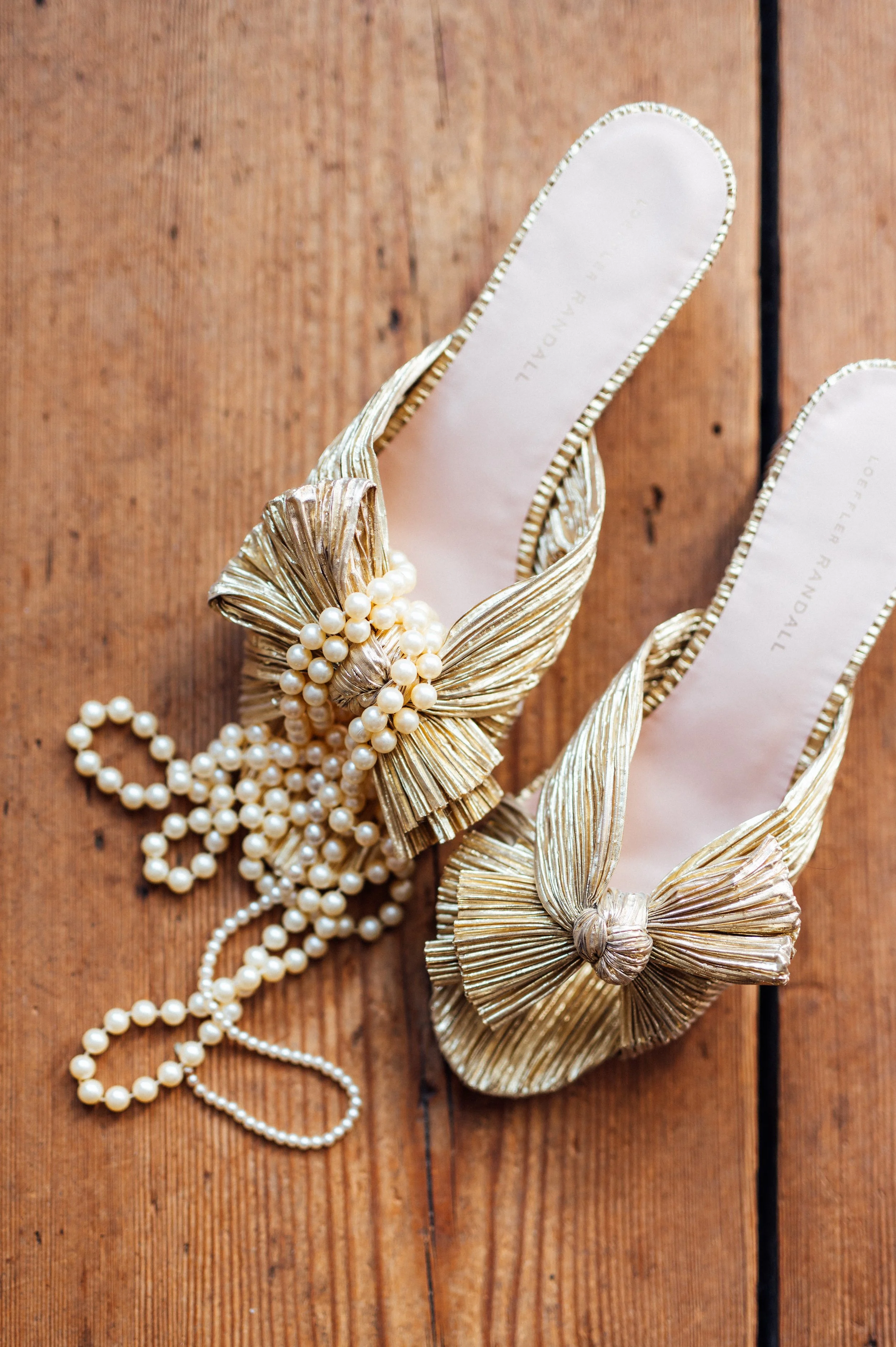 Gold high-heeled shoes with large bows and pearl embellishments, placed on a wooden surface, with a pearl necklace nearby.