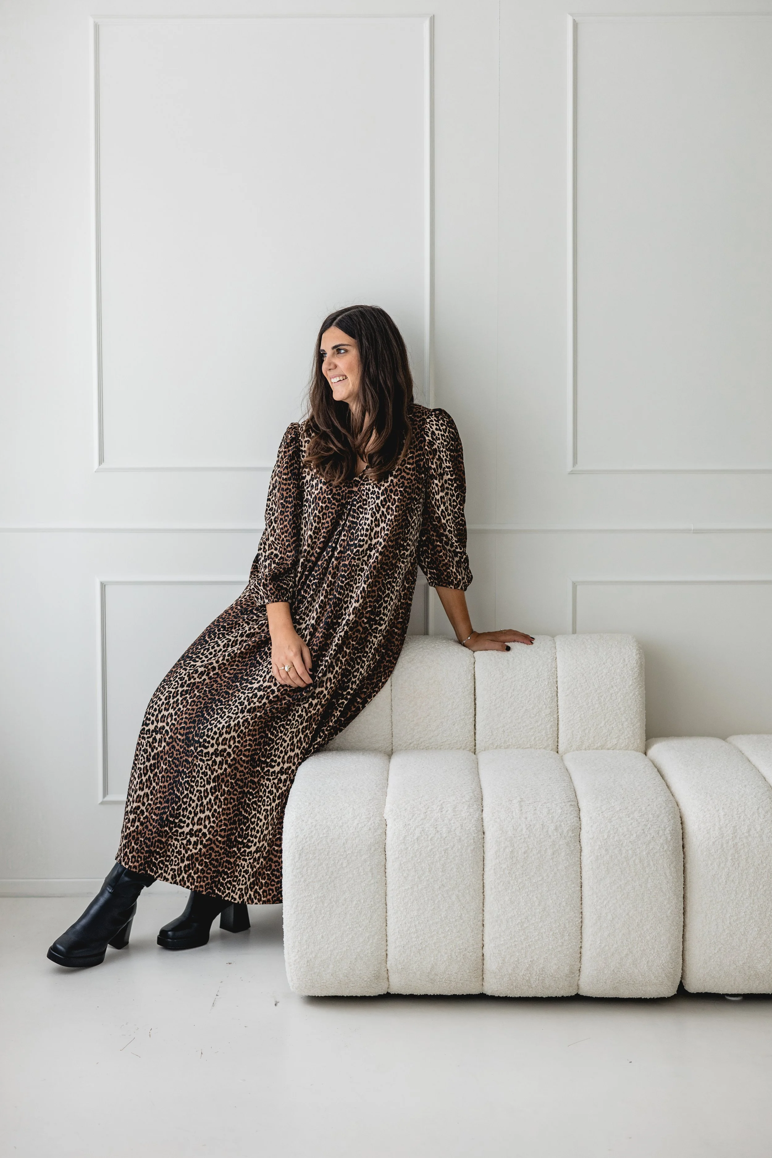 Woman wearing a leopard print dress and black boots sitting on a white textured sofa against a white wall with paneling.