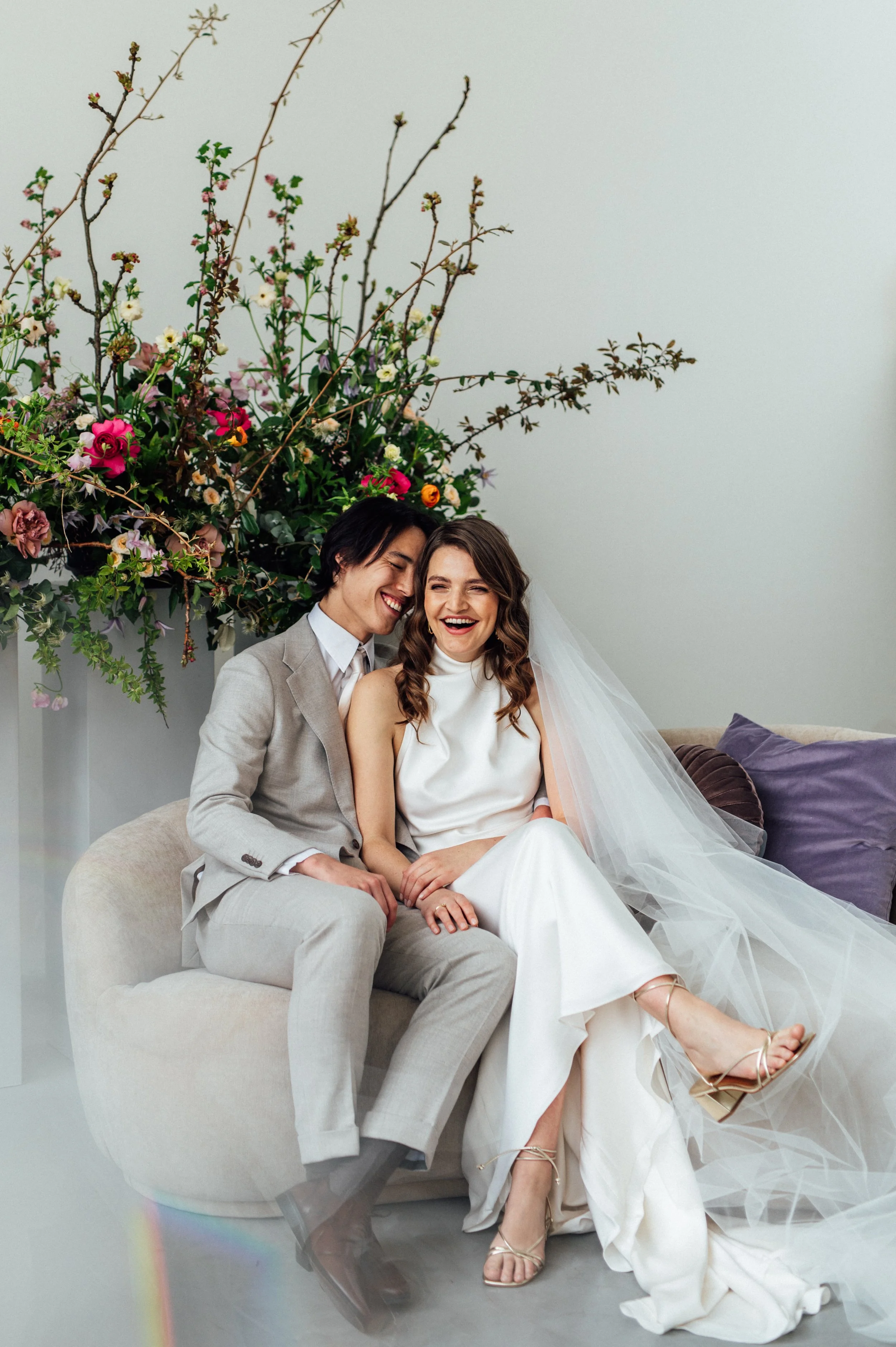 A happy couple in wedding attire sitting on a beige sofa, laughing, with a large floral arrangement behind them.