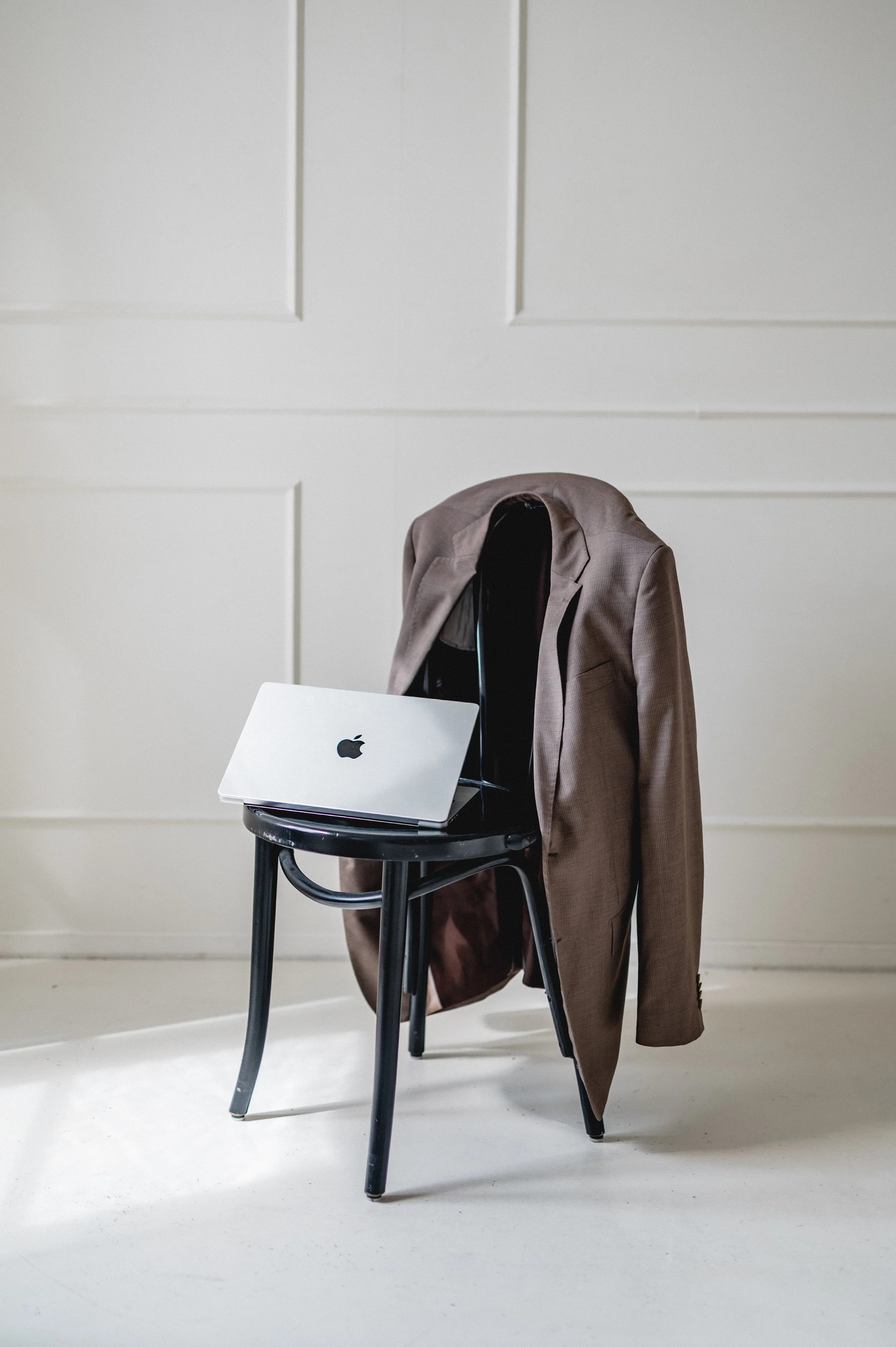 A black chair with a brown suit jacket draped over its backrest. On the chair's seat, there is a closed silver MacBook laptop. The background features a plain white wall with paneling.