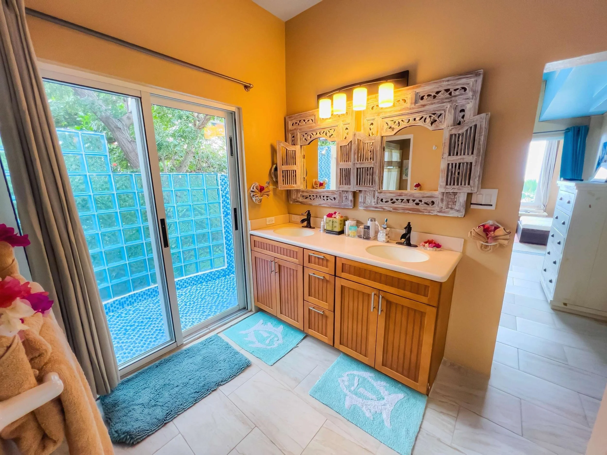 Bathroom with double sink vanity, wooden cabinet, decorative mirror with shutters, towels, and outdoor view through sliding glass door to a blue-tiled enclosed patio.