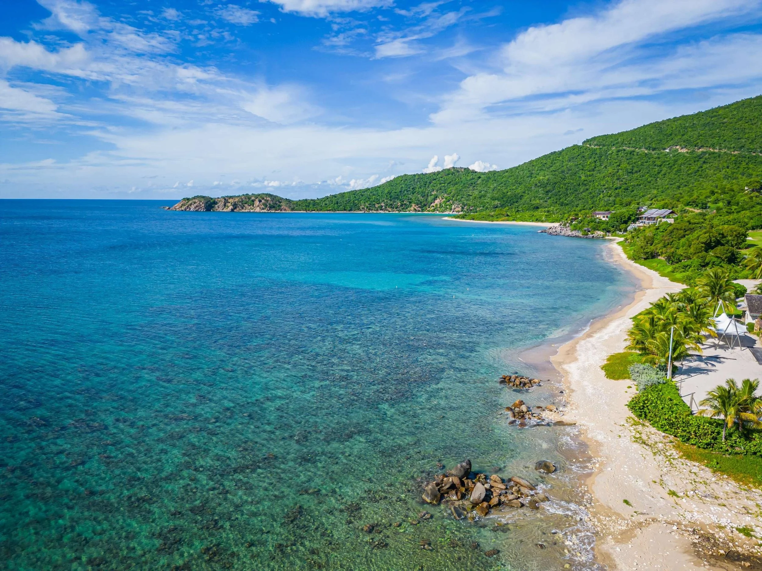 A tropical beach with clear blue water, white sandy shore, lush green hills, and palm trees.