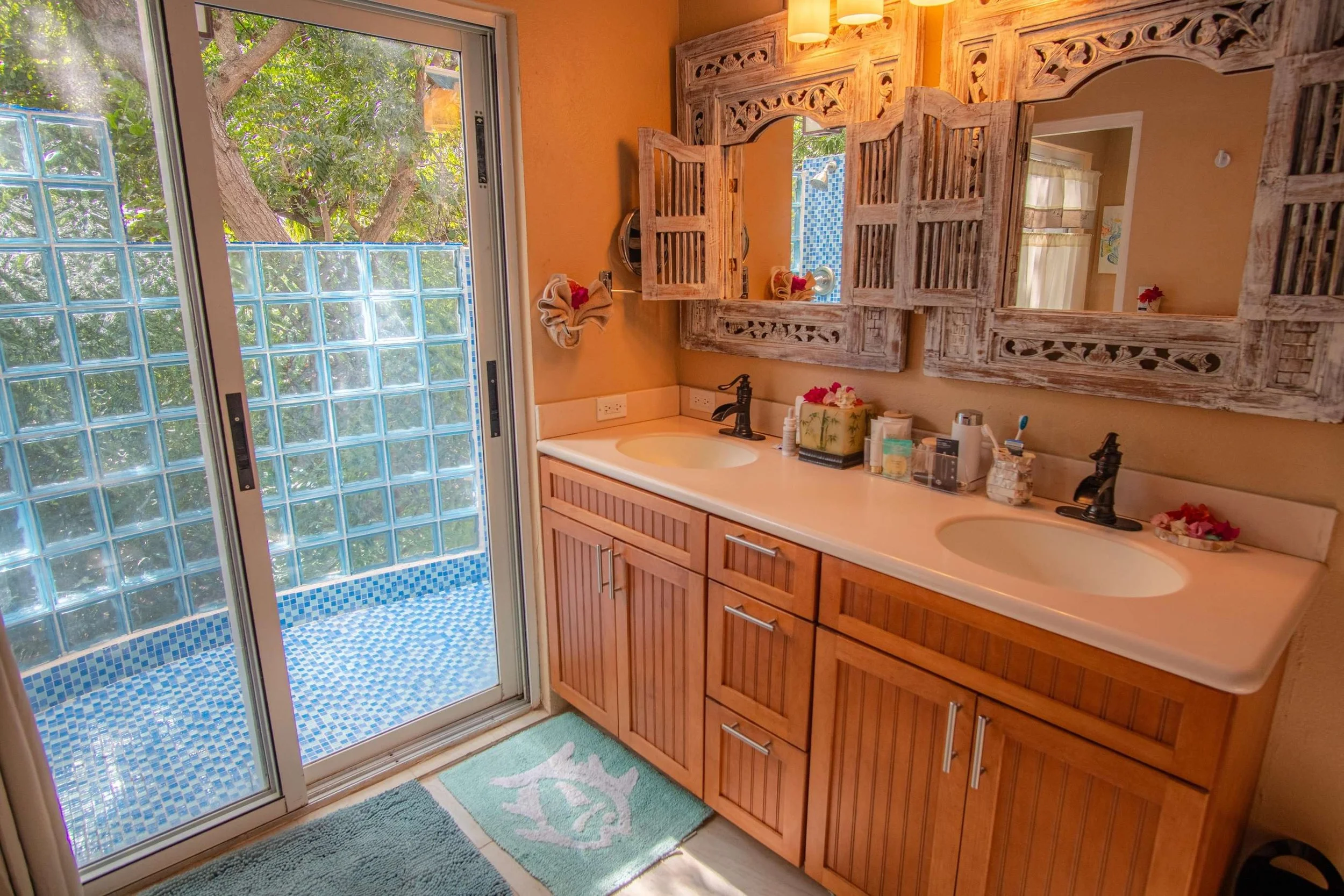 Double bathroom vanity with a large mirror, rustic wooden cabinet, two sinks, and two black faucets, decorated with small flower arrangements. A glass sliding door leads outside to a small outdoor tiled shower area with blue tile mosaic and glass blo