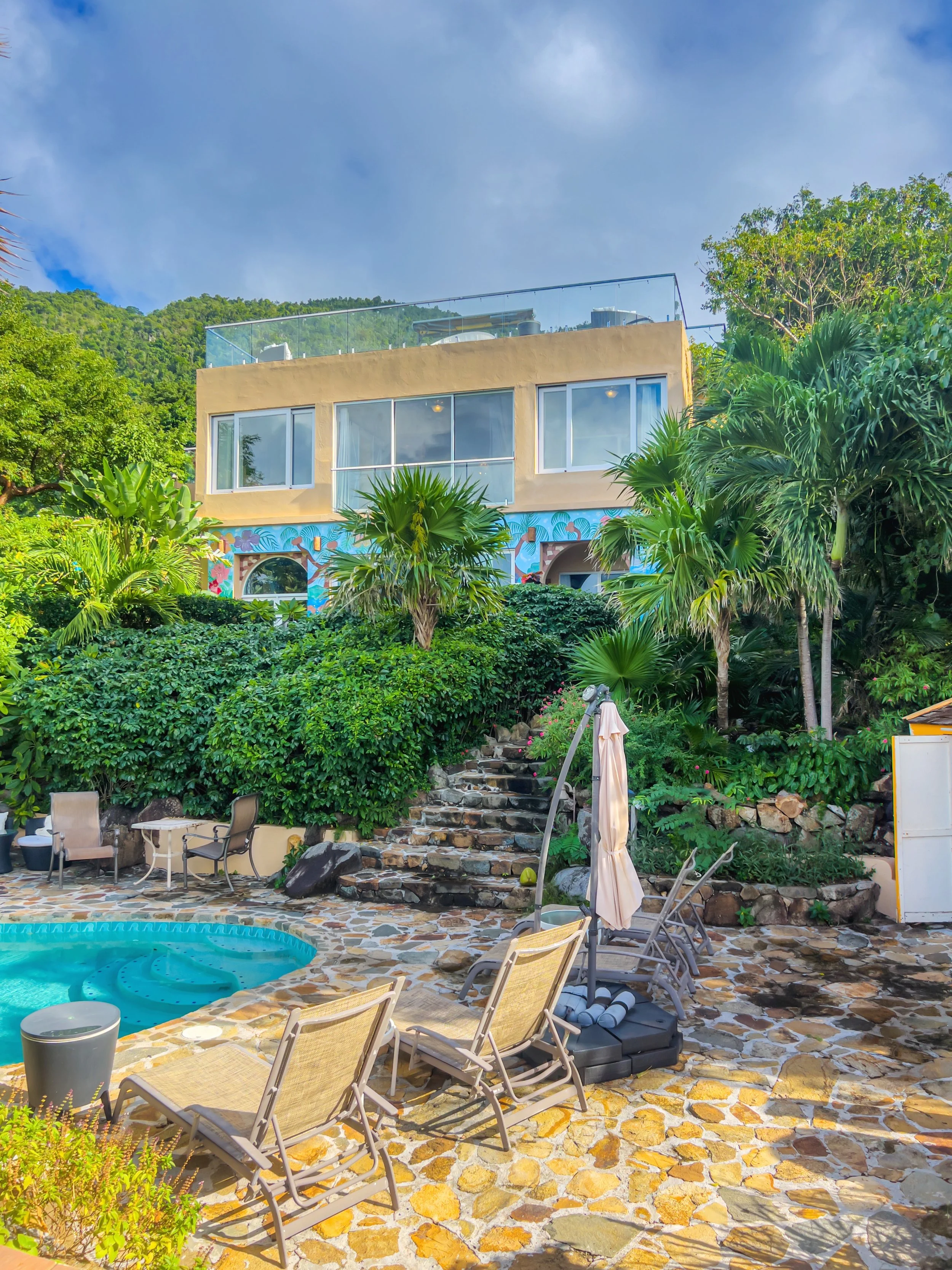 Outdoor pool area with lounge chairs, umbrellas, and a stone patio, with lush green plants and a multi-story house in the background under a partly cloudy sky.