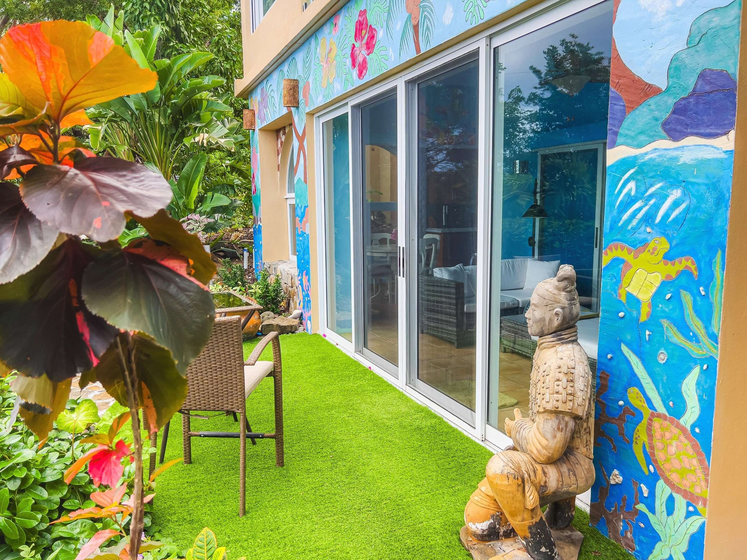 Outdoor patio with green artificial turf, a wicker chair with cushioned seat, colorful tropical tattoos on the wall, lush green plants, and a carved wooden Buddha statue. The patio is enclosed by sliding glass doors, revealing an interior with white 