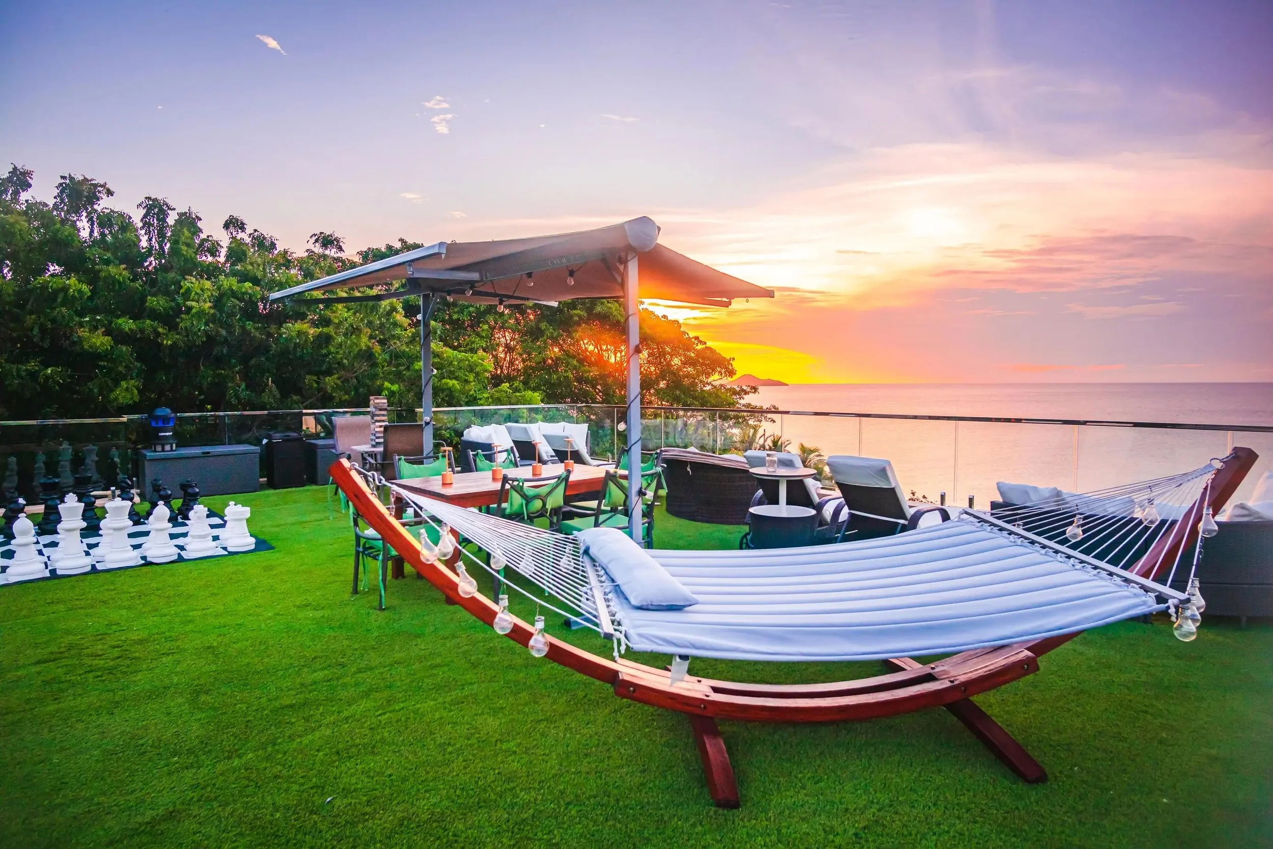 Outdoor rooftop patio with hammock, lounge chairs, dining table, and chess set overlooking ocean at sunset.