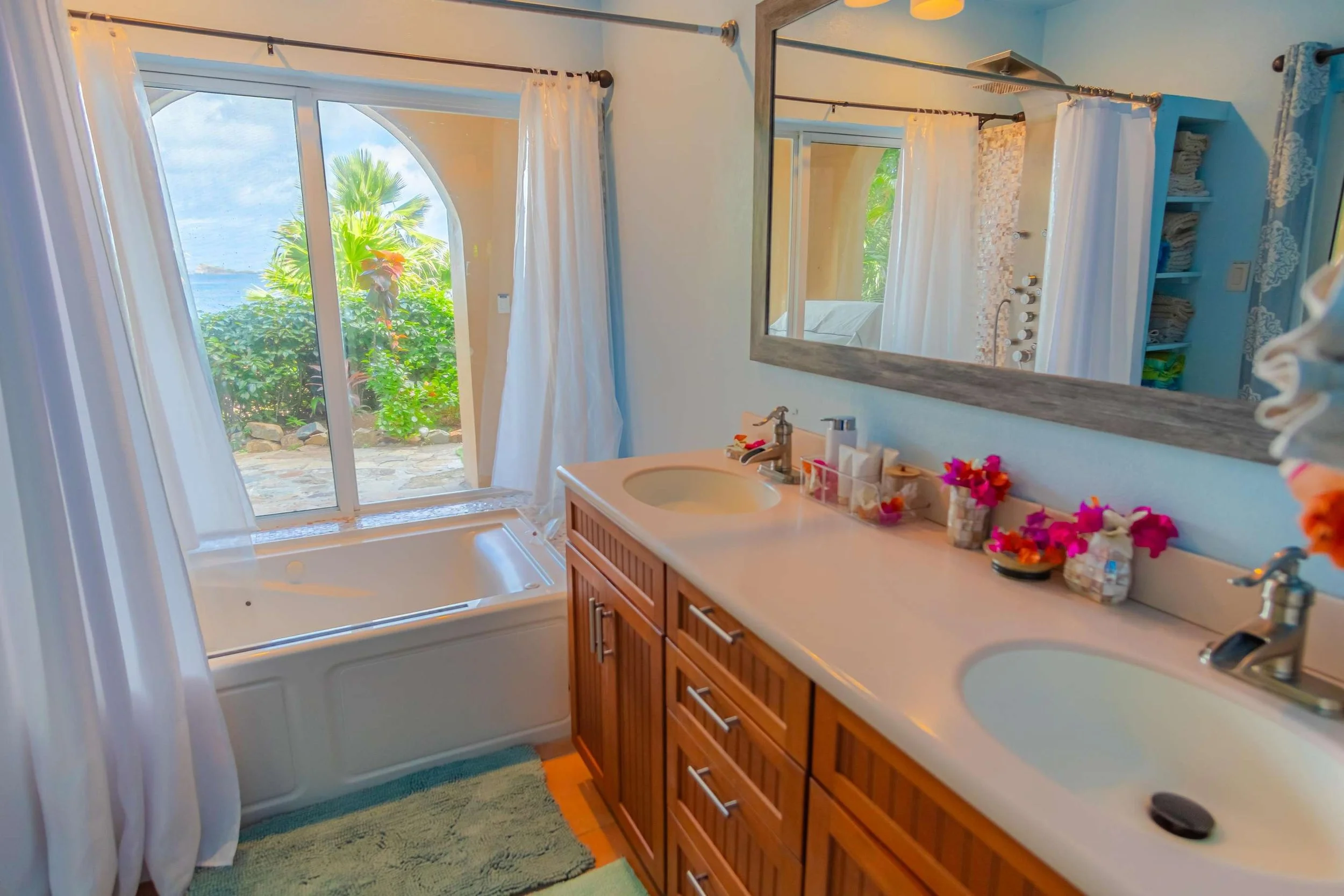 A bathroom with dual sinks, a large mirror, and a window with a view of tropical greenery and a pathway outside. The counters are decorated with flowers, and there's a bathtub near the window with white curtains.