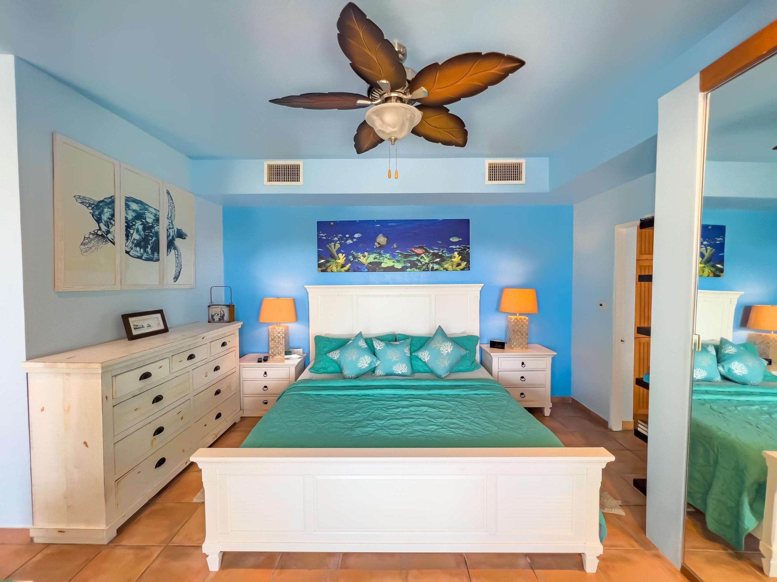 A bedroom with a tropical theme featuring blue walls, a white bed frame, turquoise bedding, and pillows with coral designs. Two orange lamps are on white nightstands, and there is a fish-themed painting above the bed. A mirrored closet door reflects 