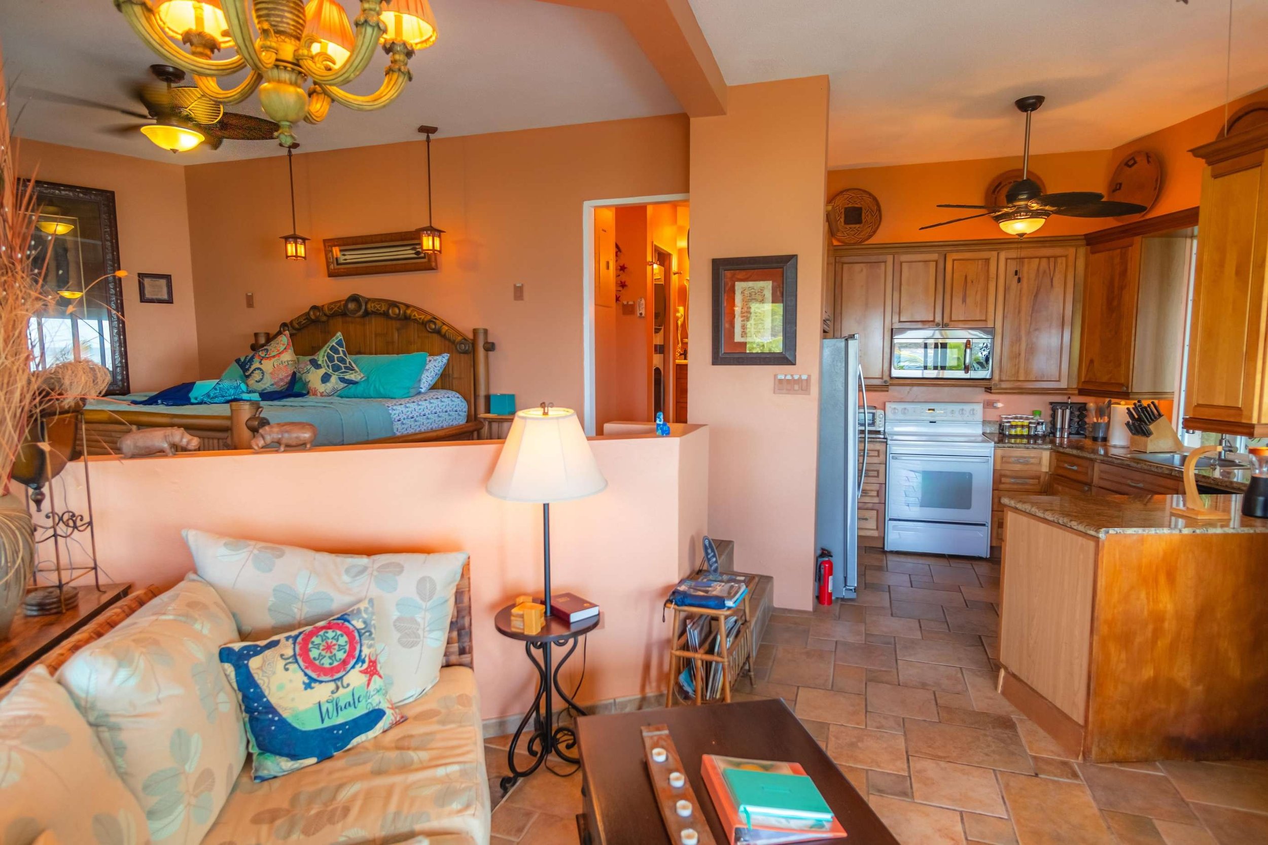 A cozy living space with a glimpse of the kitchen. The living area has a floral-patterned sofa with a decorative pillow, a side table with a lamp, and a coffee table with notebooks. The kitchen features wooden cabinets, a white stove, a microwave, an