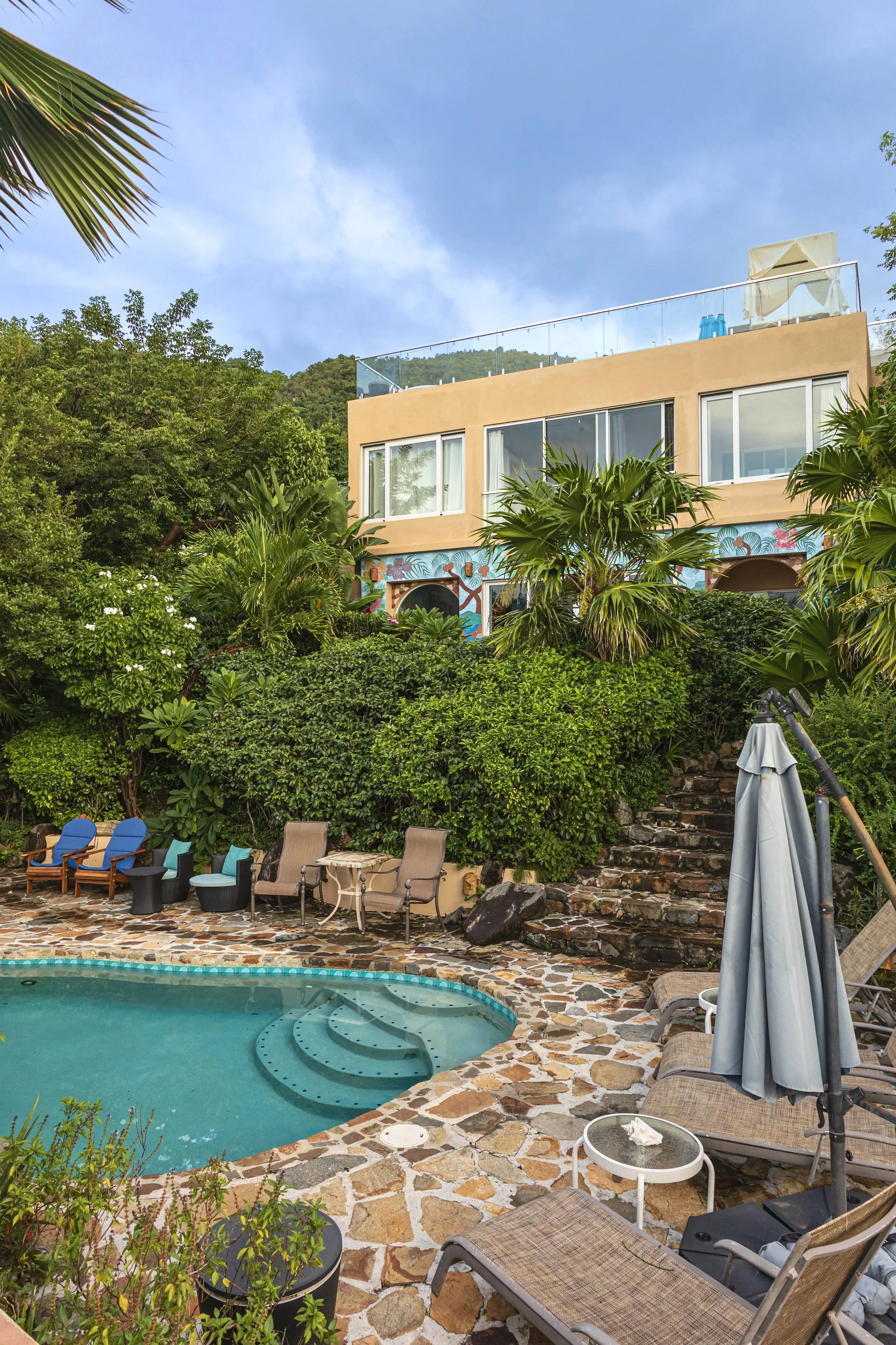 A swimming pool with a stone deck surrounded by lounge chairs and lush green plants, with a multi-story building and a scenic hillside in the background.