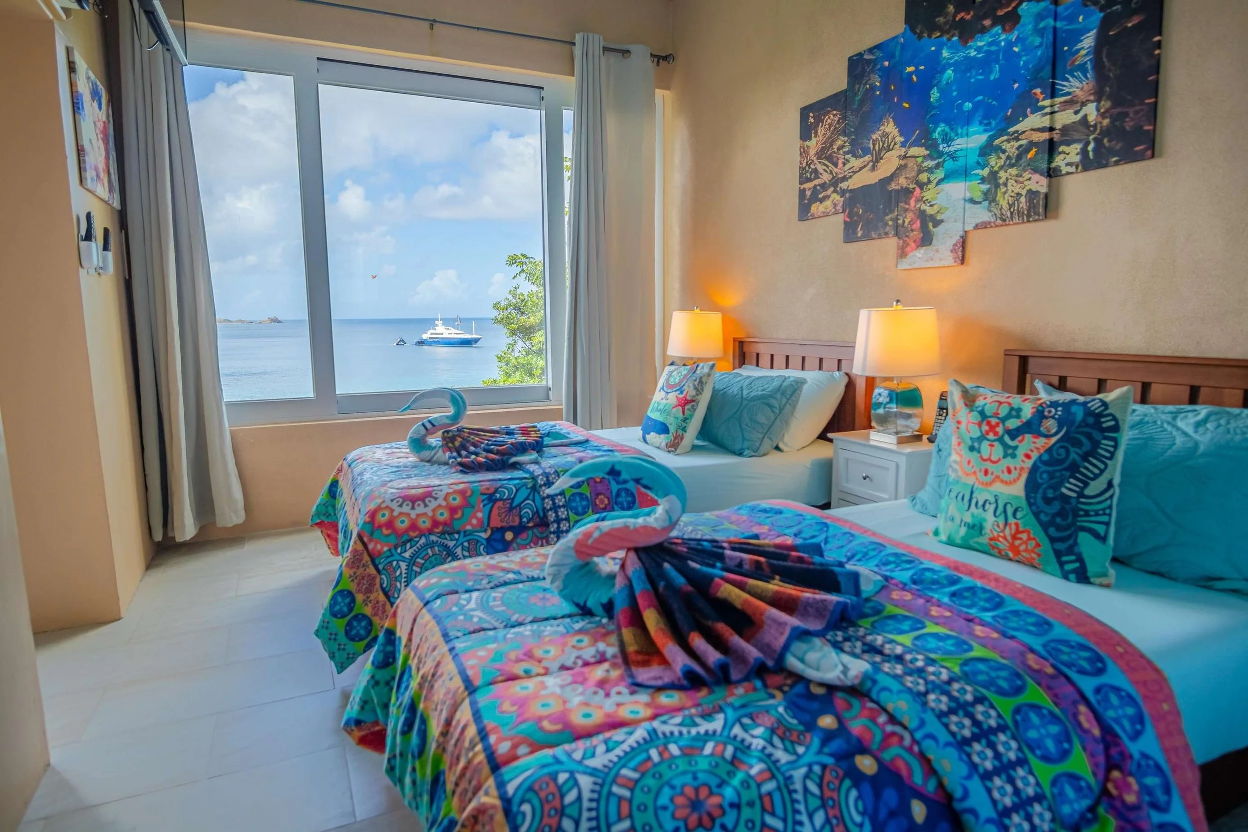 Beachfront hotel room with two beds, colorful bedding, and towels shaped like swans, overlooking the ocean and a yacht through large window.
