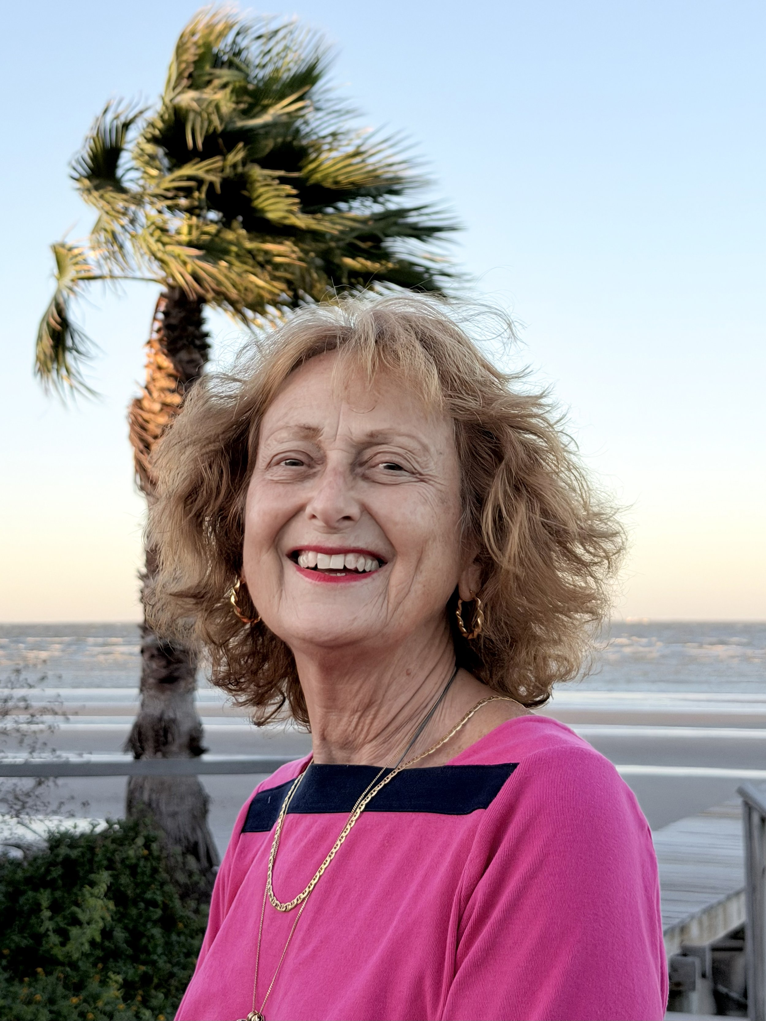 Portrait of a smiling woman with wavy blonde hair, wearing a pink top with black accents, gold jewelry, and standing outdoors near a beach with a palm tree in the background during sunset.