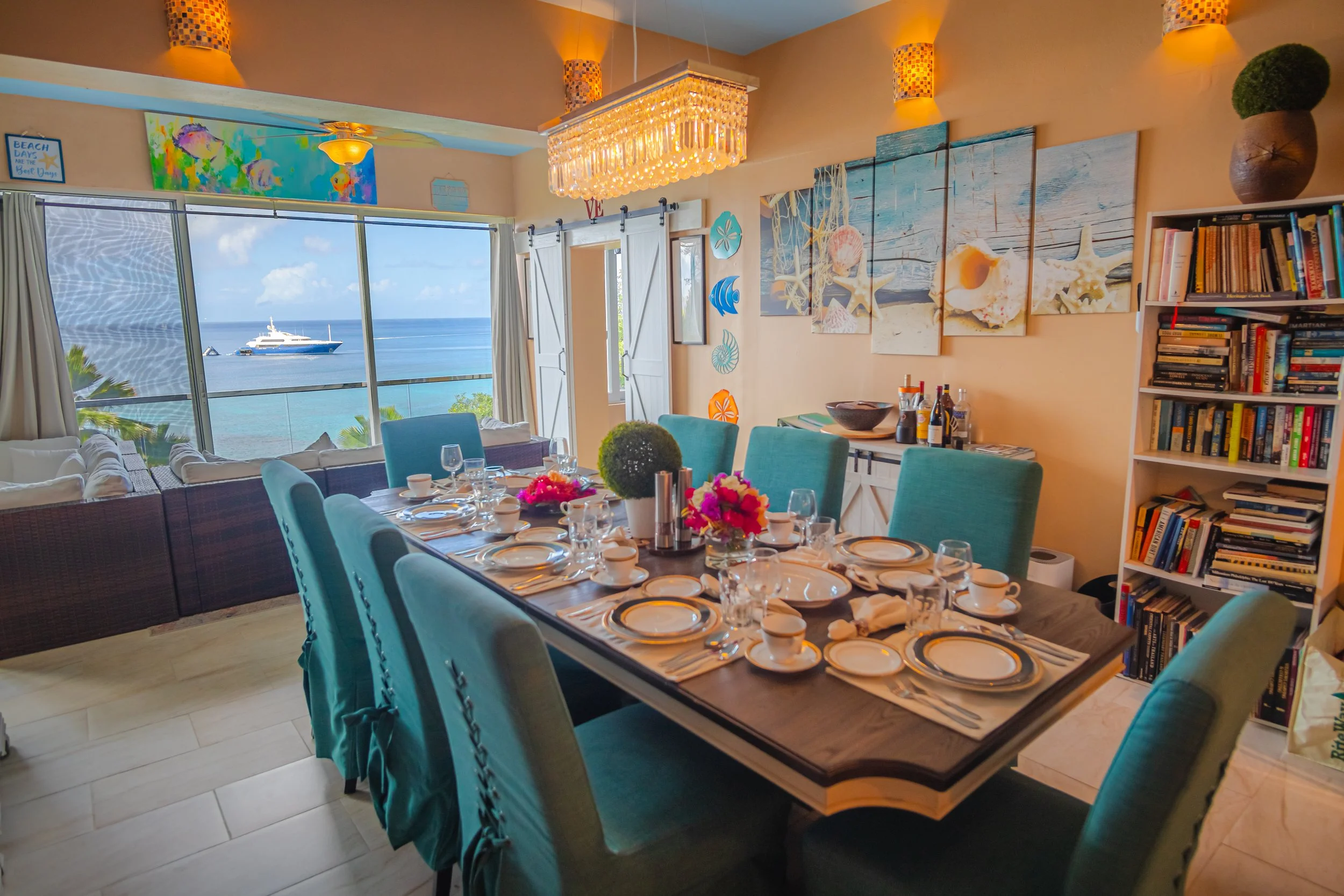 Beautiful tropical dining room with ocean view, decorated with beach-themed art, a chandelier, and a table set for a meal with flowers.