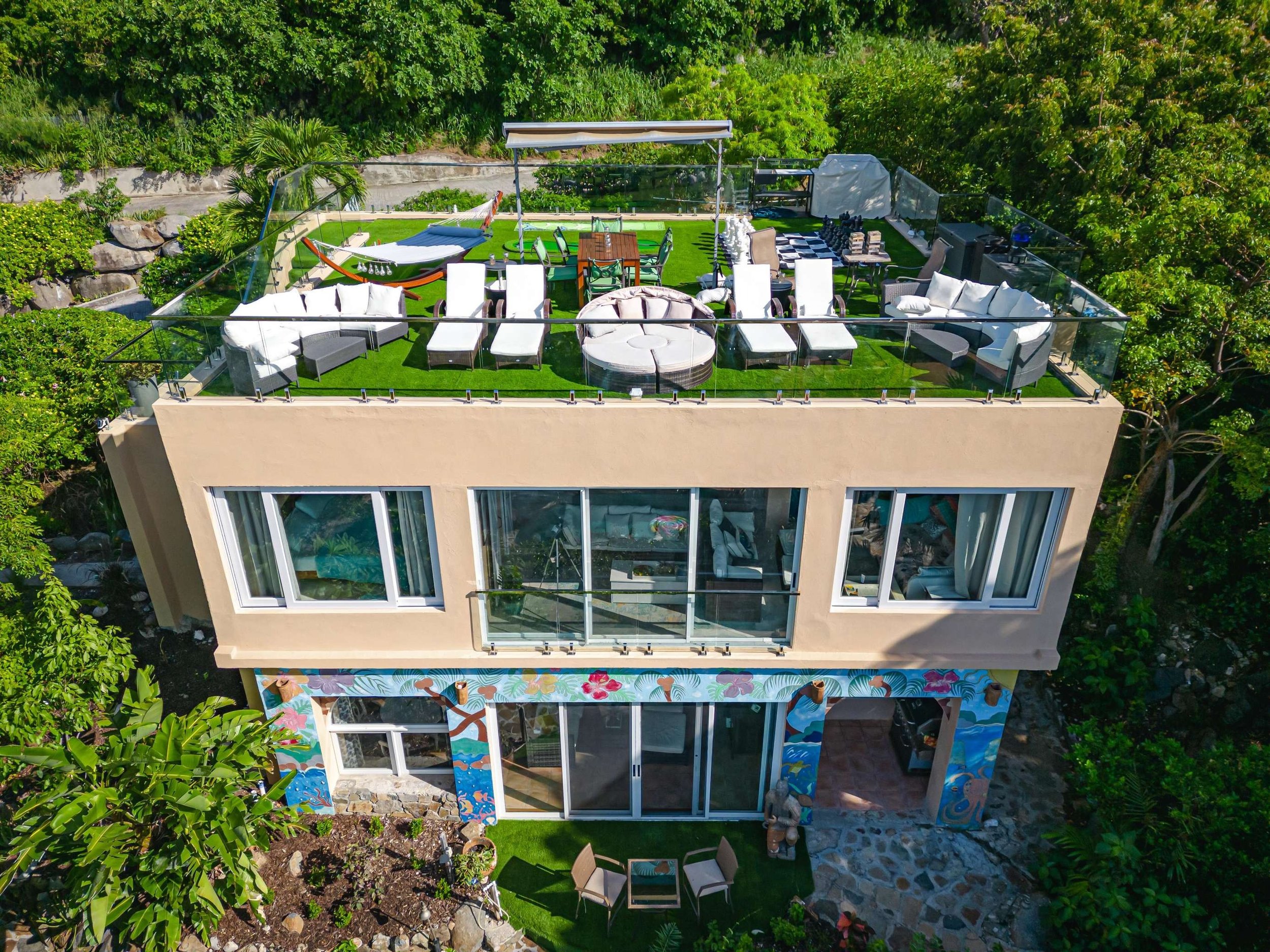 A modern, multi-story house with a rooftop terrace surrounded by green trees and plants. The rooftop has outdoor seating, a hammock, a dining table, and lounge chairs, all on artificial grass, with a glass railing.