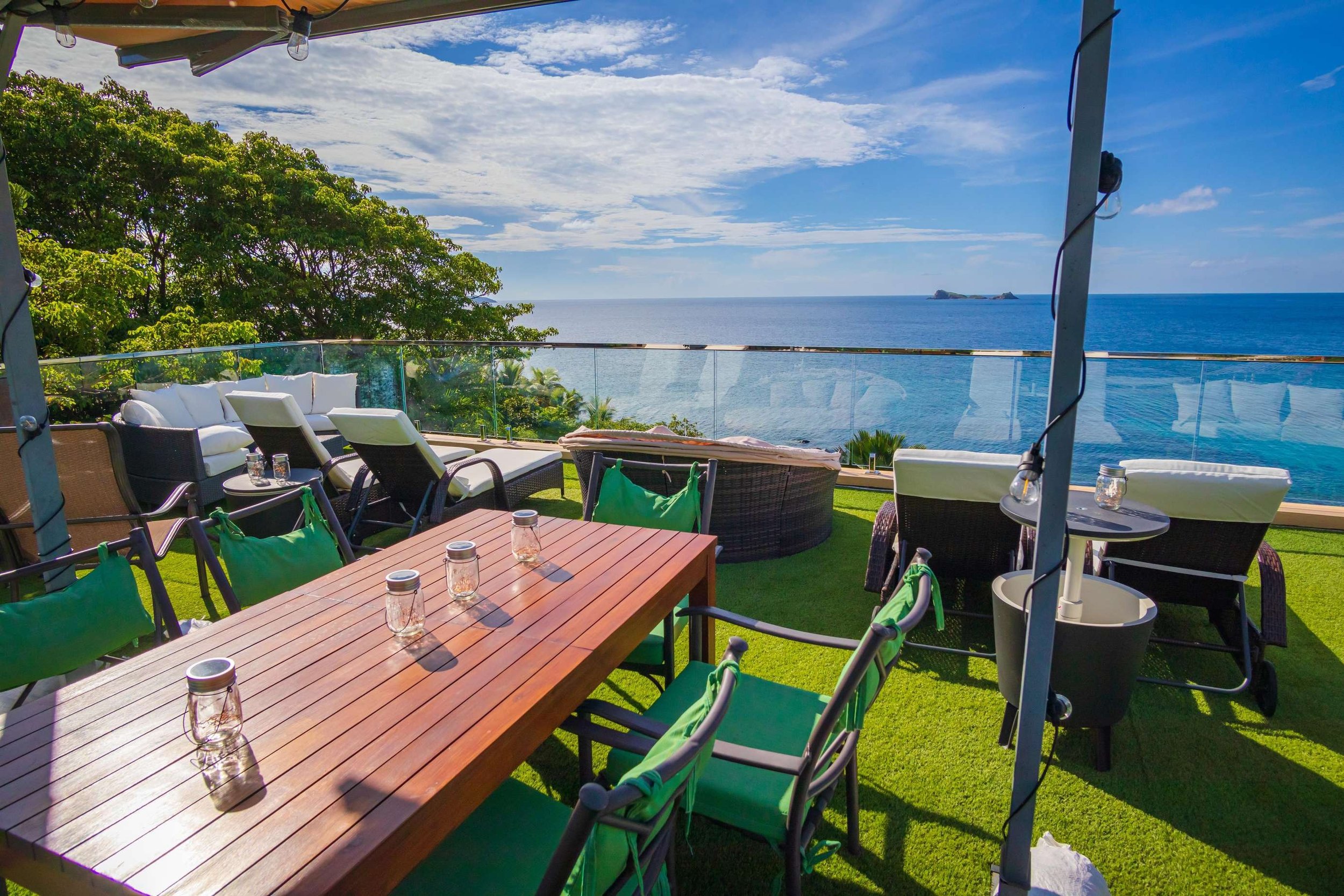 Outdoor rooftop patio with green chairs, a wooden table, and a view of the ocean and blue sky with some clouds.