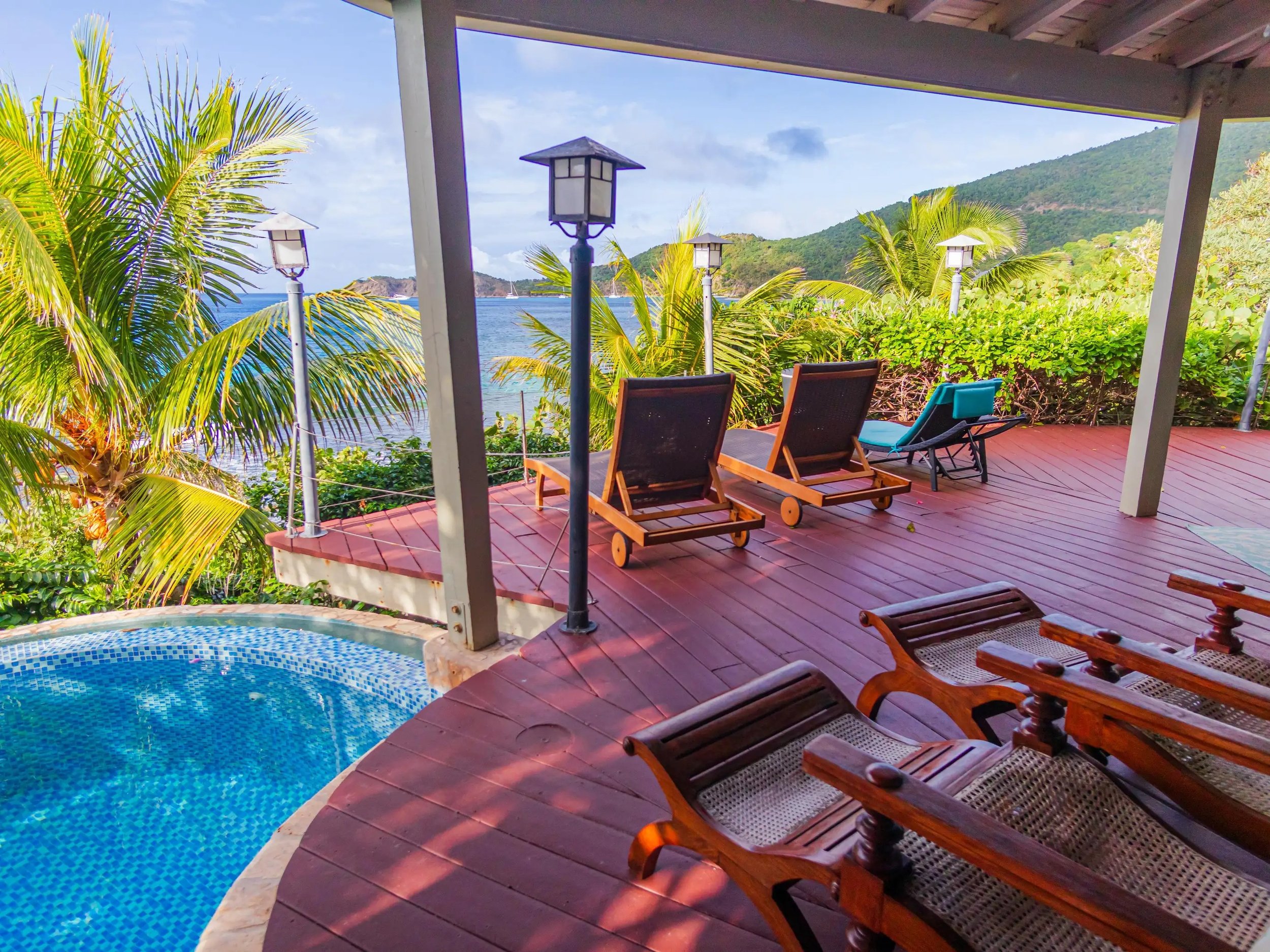 A tropical patio with a swimming pool, wooden lounge chairs, and views of a lush green hillside, palm trees, and the ocean.