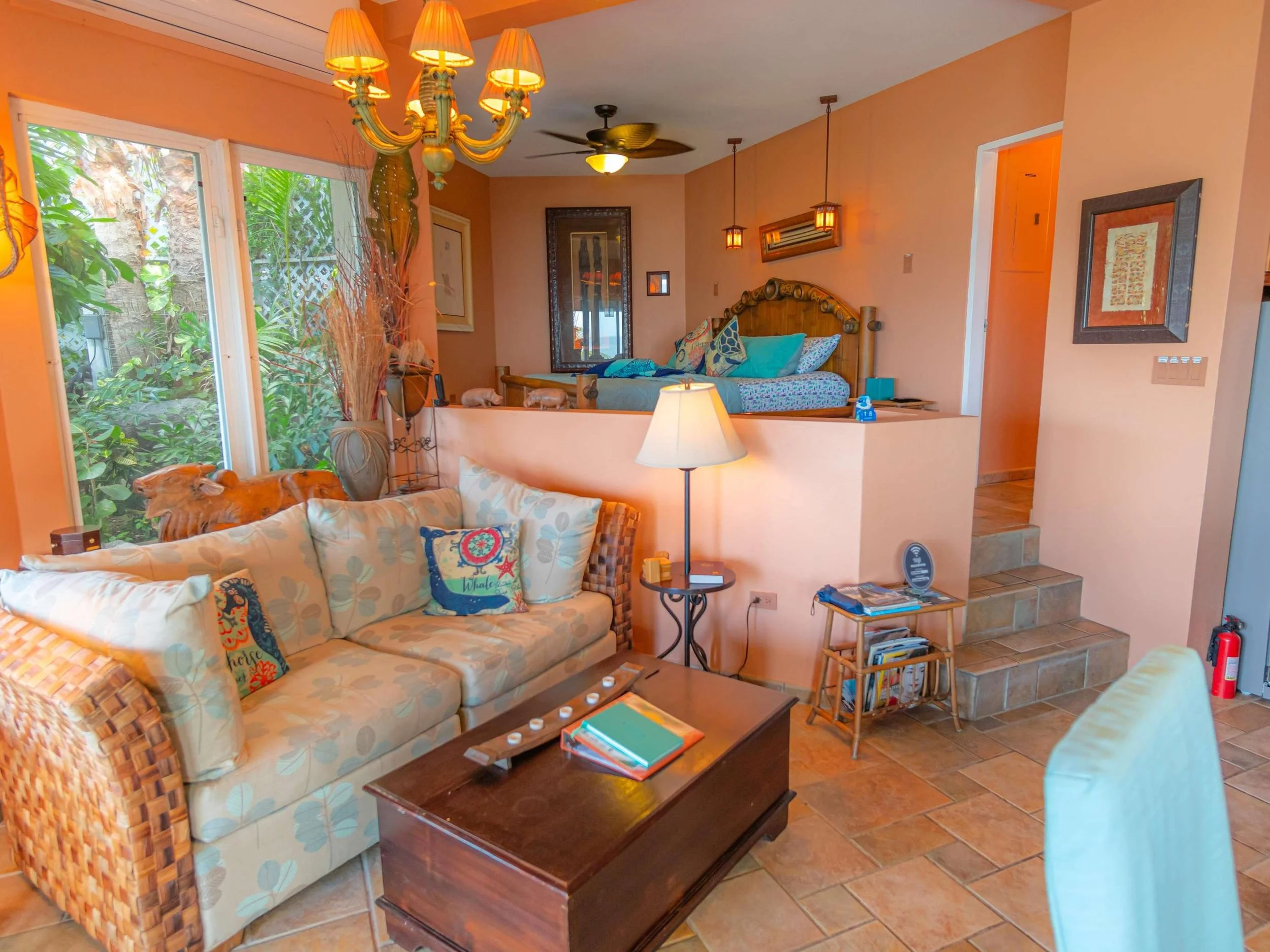 Living room with a floral-patterned sofa, throw pillows, a wooden coffee table, and a lit floor lamp. There are large windows with greenery outside, a chandelier hanging from the ceiling, and a partially raised bedroom area with a bed and pillows. Th