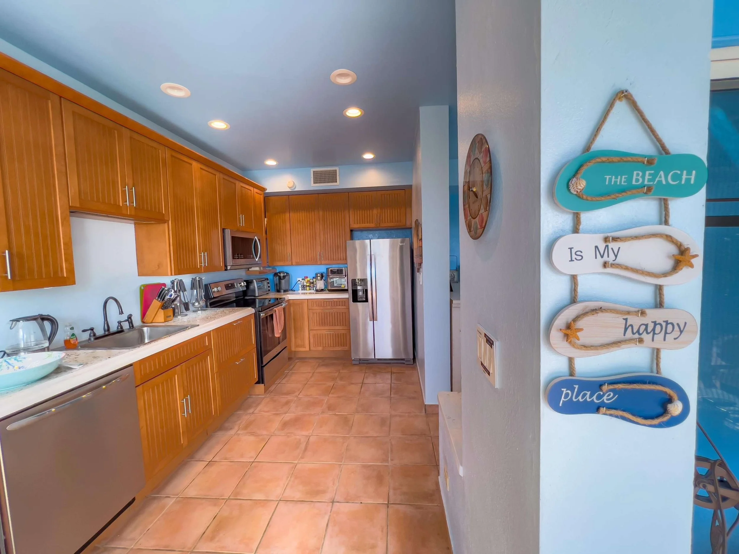 Kitchen with wooden cabinets, stainless steel appliances, tiled floor, and a decorative sign on the wall that reads "The Beach Is My Happy Place."