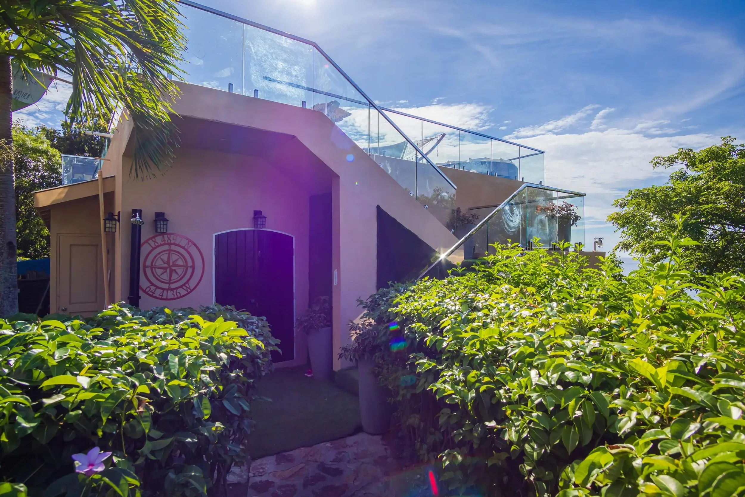 A building with pink walls and a black door, surrounded by lush green bushes and plants, with a glass fence on a balcony above, under a bright blue sky with some clouds.