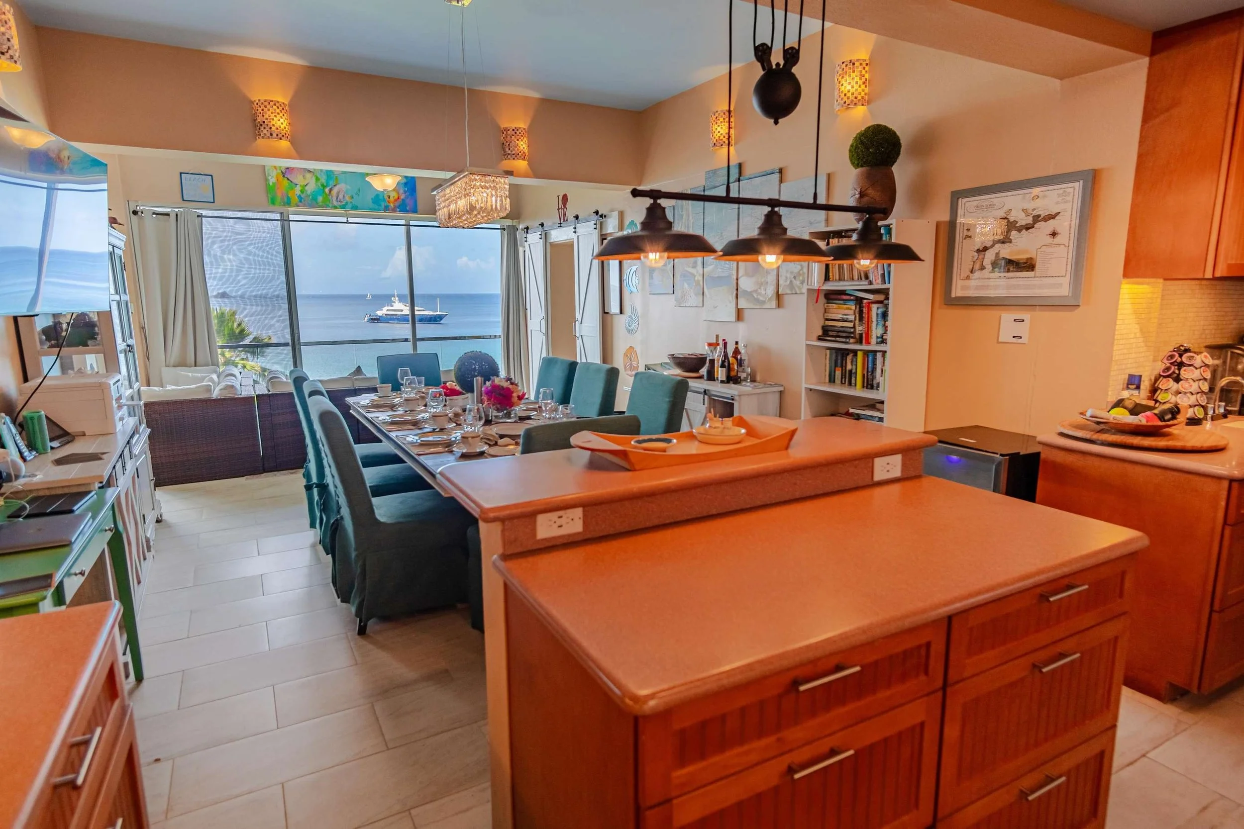 Open-concept living and dining area with a large window view of the ocean, a yacht, and a sailboat, featuring a dining table set for a meal, a kitchen island, bookshelves, and cozy seating.